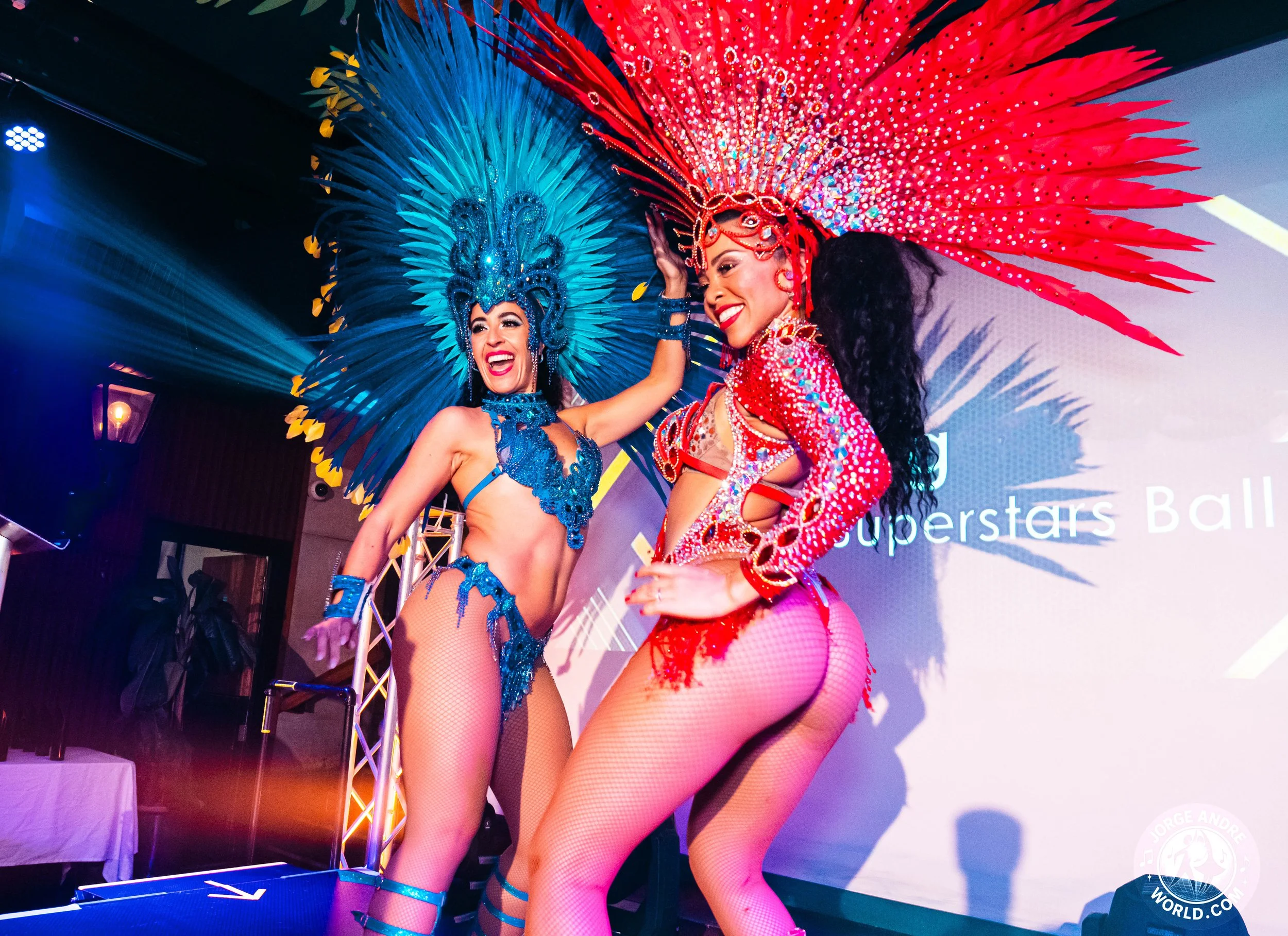 Two women in elaborate carnival costumes with big feathered headdresses, performing on stage at an event.