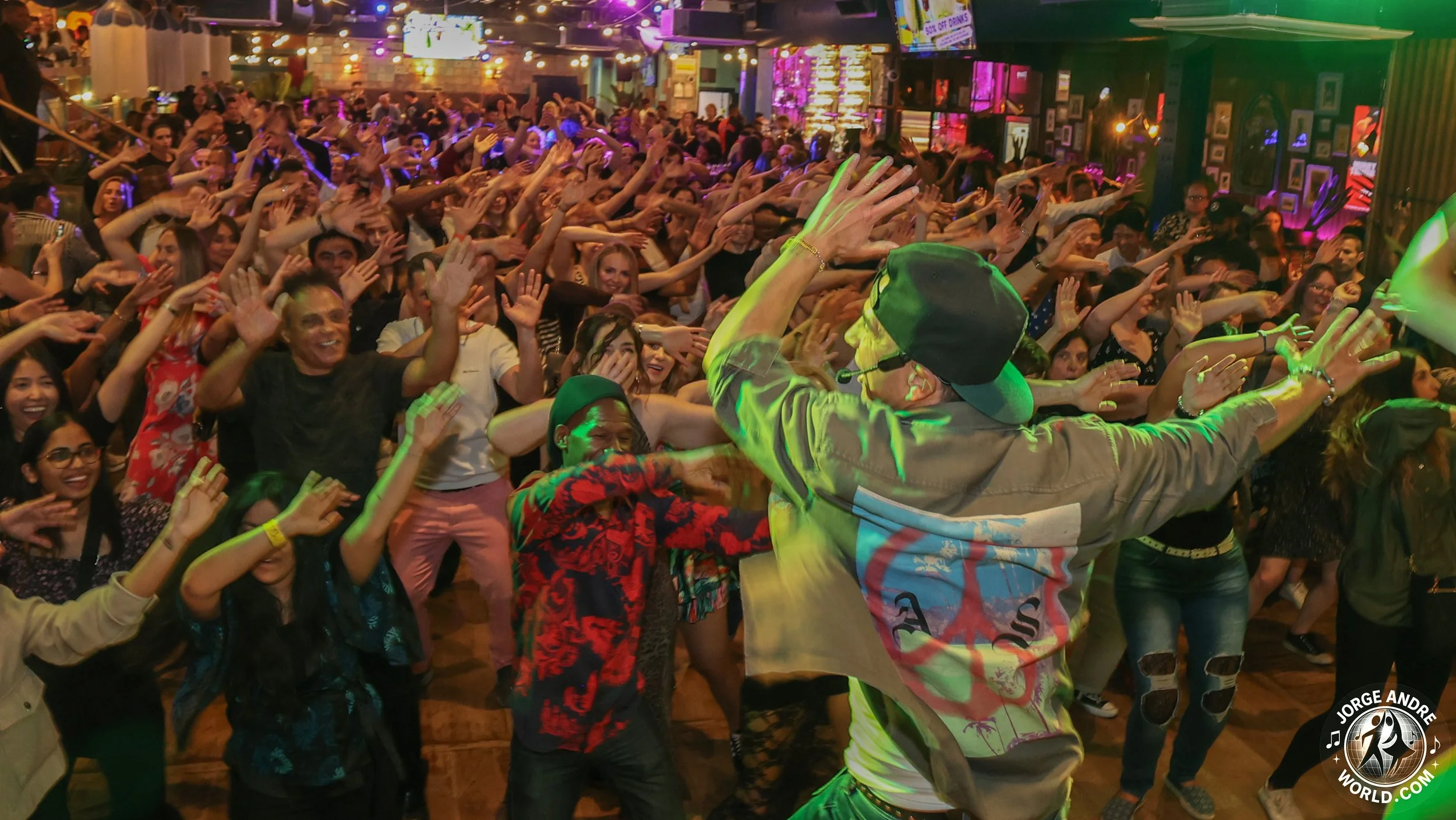 A lively indoor concert or event with a large crowd of people dancing and waving their hands in the air, with a performer or host in the foreground engaging with the audience.