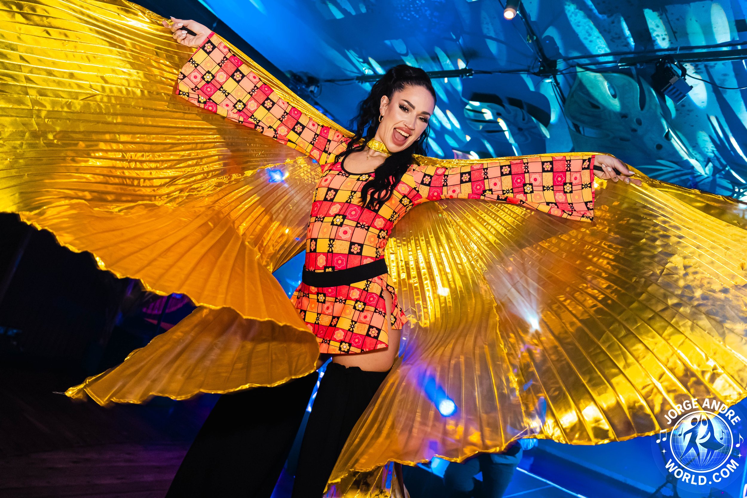 A woman dancing with large golden wings in a vibrant environment, wearing a red and yellow patterned outfit with flared sleeves. She has black hair, and an energetic expression, with a blue background and stage lighting.