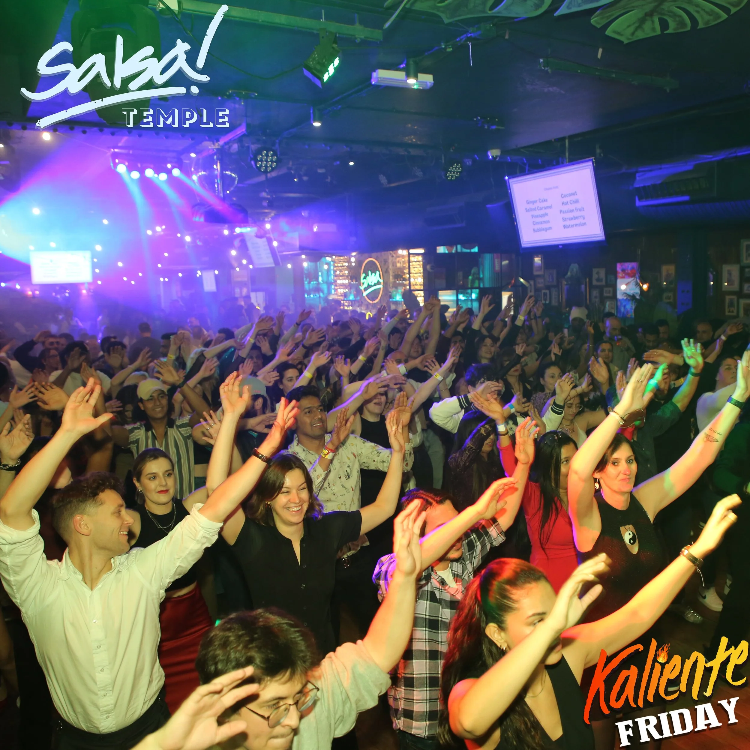 Crowd of people dancing with their hands raised in a nightclub or concert venue with colorful lighting and a neon sign that says 'Salsa'.