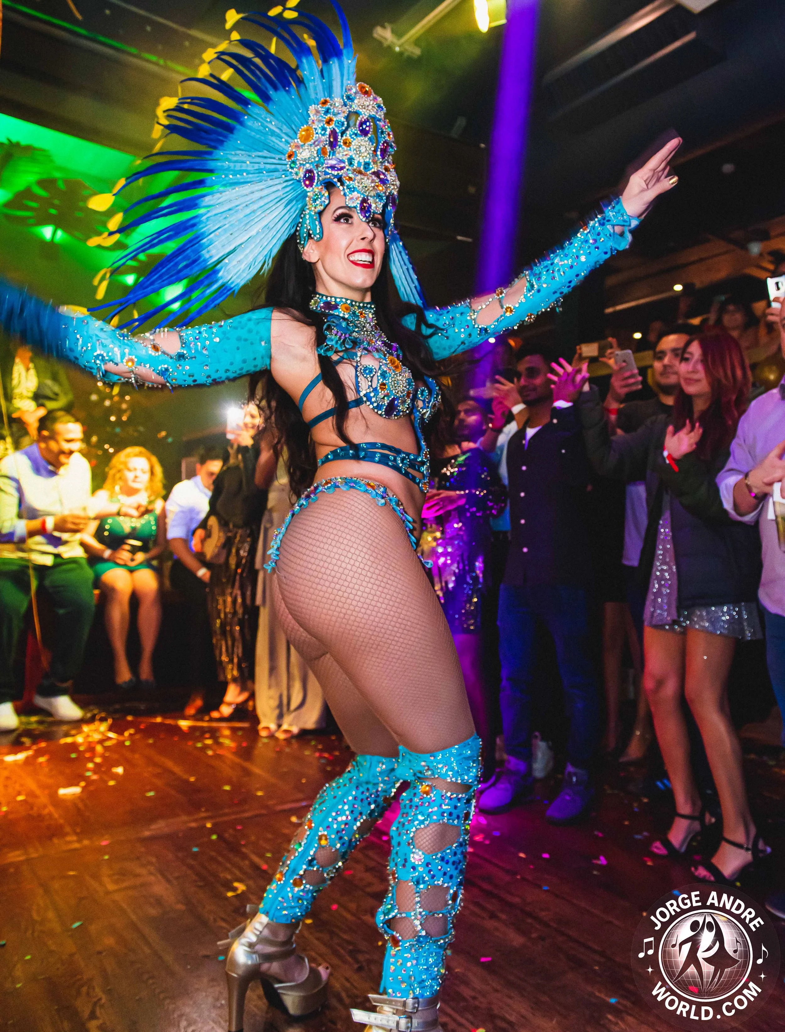 A performer in a vibrant blue costume with large feathered headdress and rhinestone embellishments dances in a crowded indoor celebration.