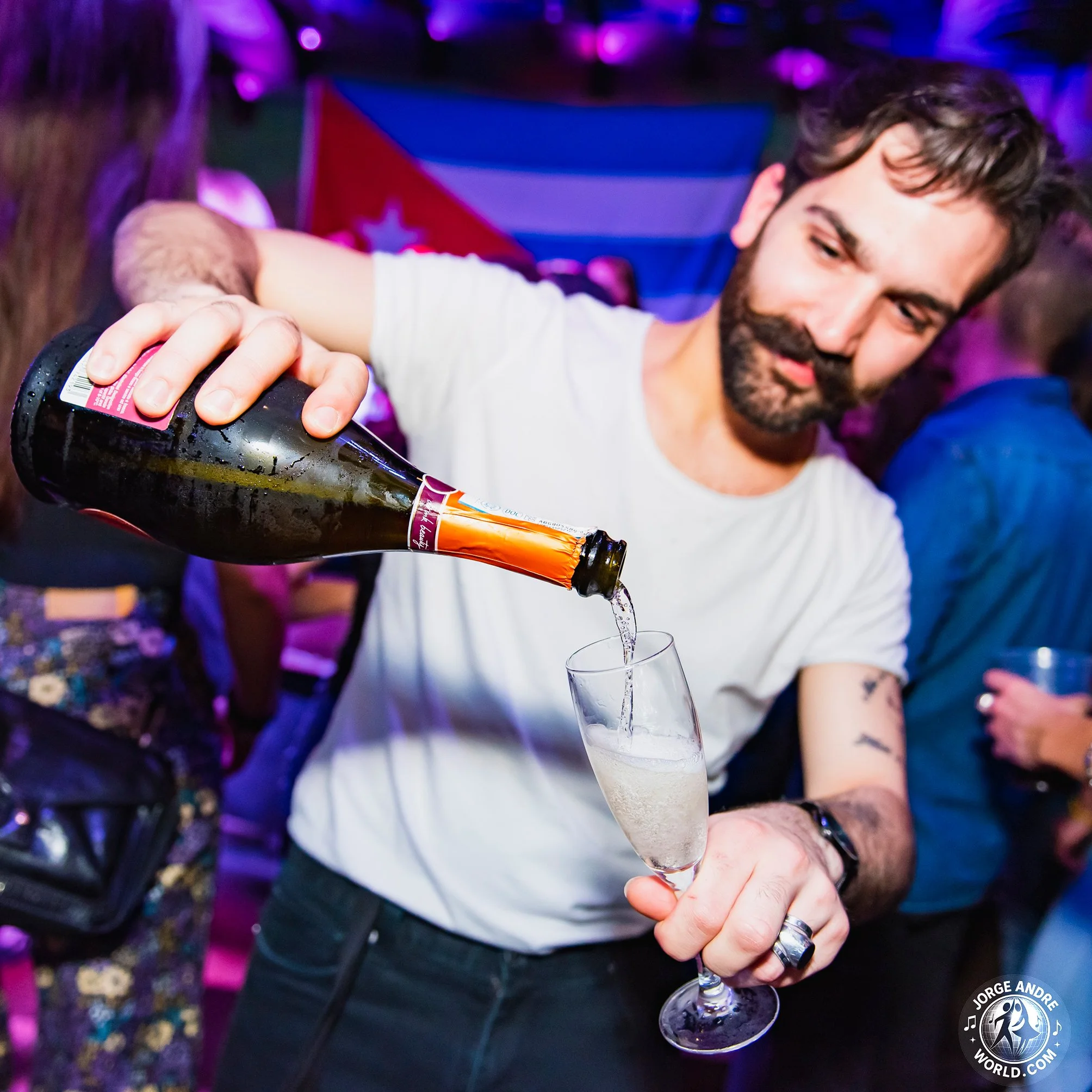 A man with dark hair and a beard pours champagne into a flute at a celebration event, with colorful lighting and a person wearing a flag in the background.
