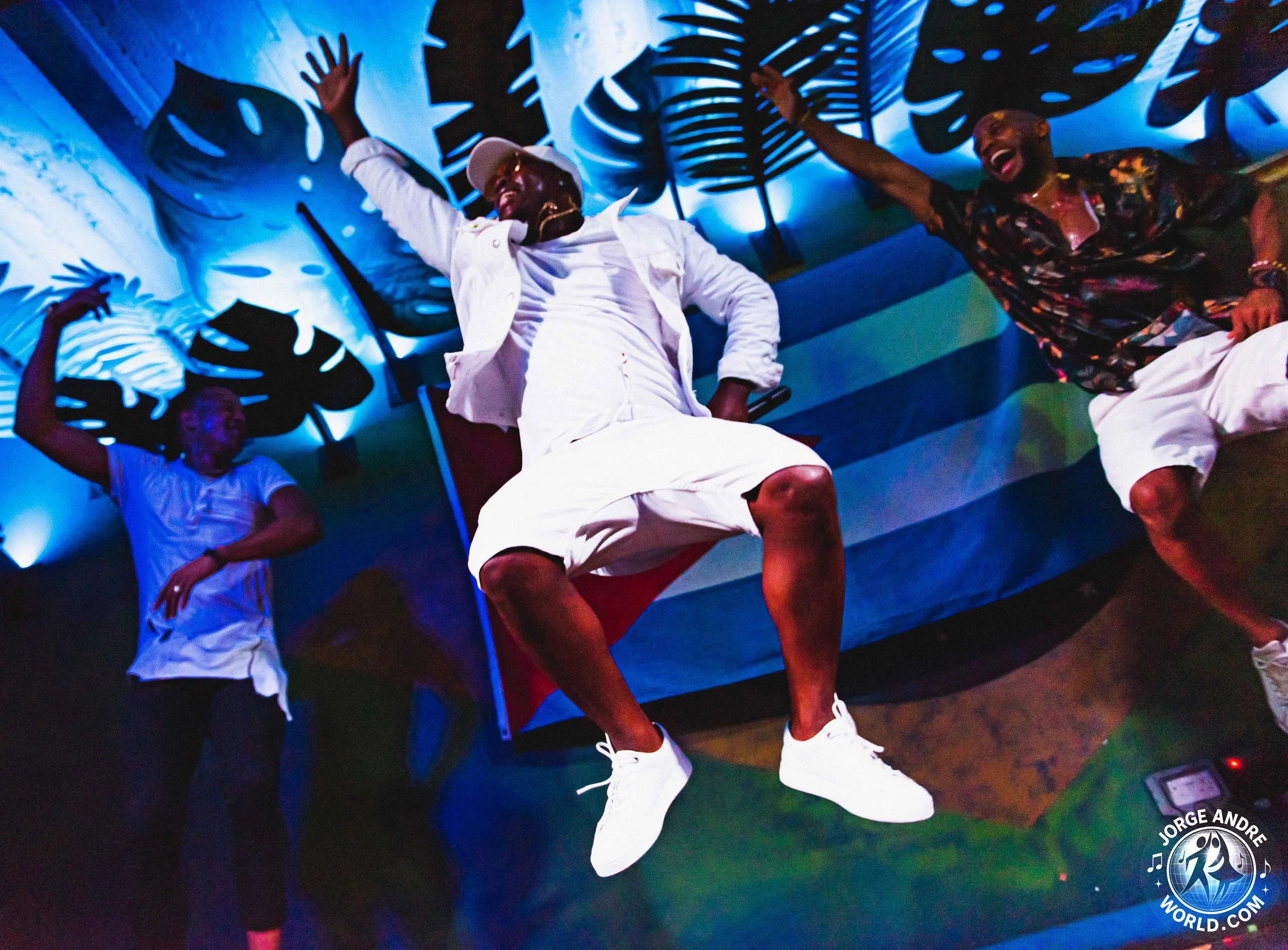 Four people dancing and having fun indoors with a blue leafy mural background, colorful lighting, and a stage. The person in the center is jumping with a white outfit and white shoes, smiling. The others are sitting or standing around, also smiling a
