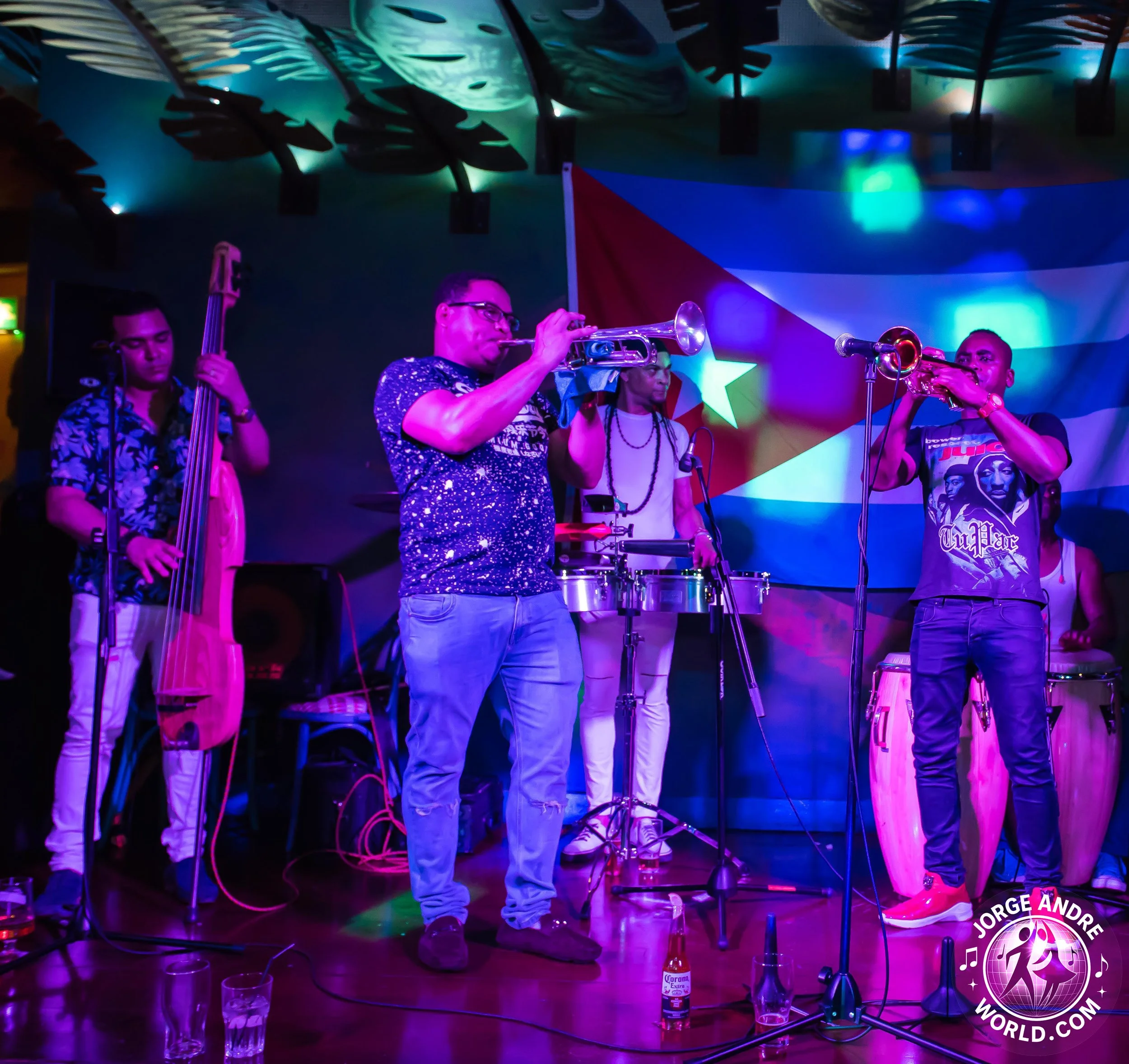 A band performing on stage with a Puerto Rican flag in the background. The band includes a double bassist, a trumpet player, a percussionist, and a conga player, with colorful stage lighting and glasses of drinks in the foreground.