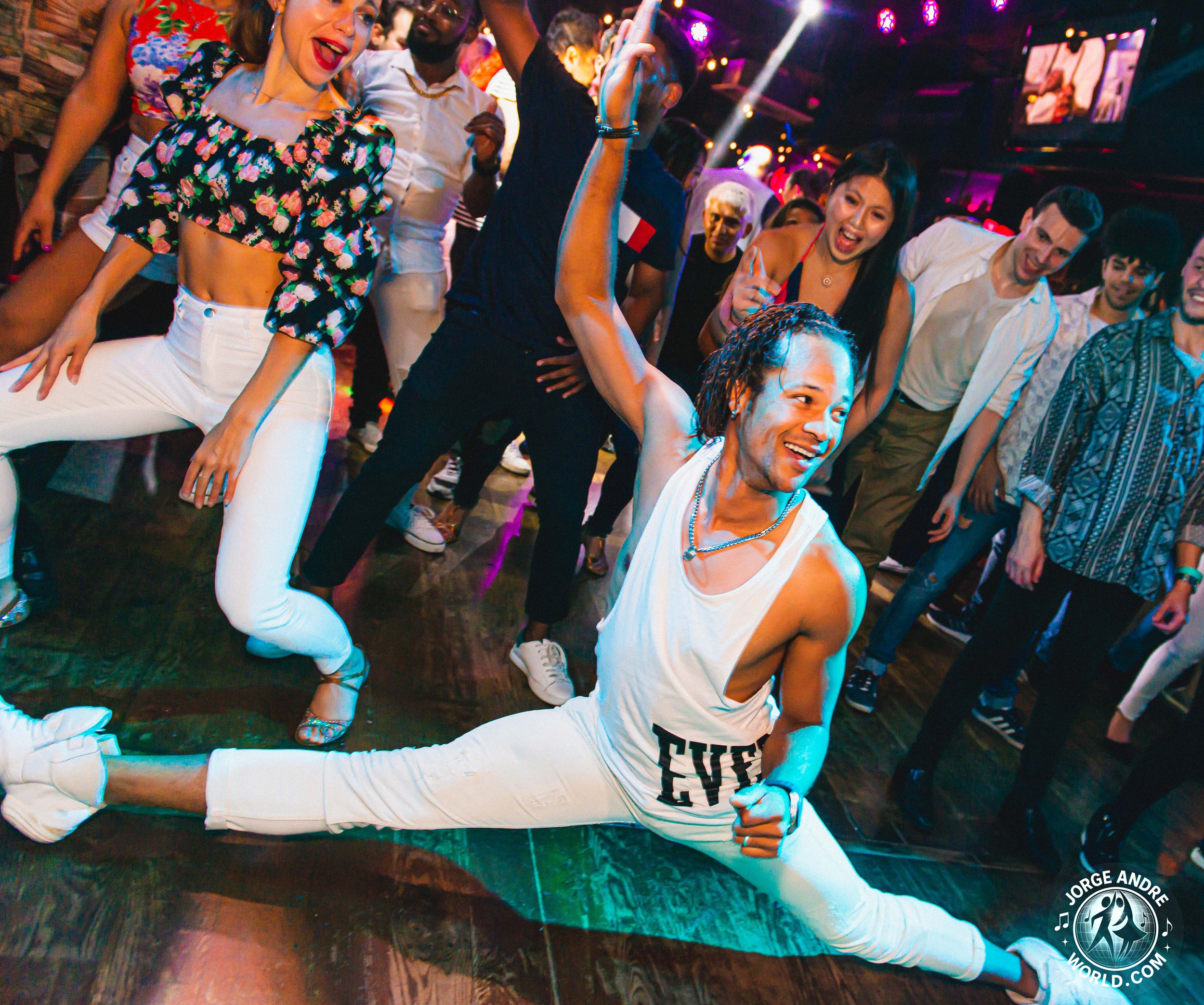 People dancing at a lively party or nightclub, with a man doing a split on the floor in the foreground and others dancing and enjoying themselves around him.