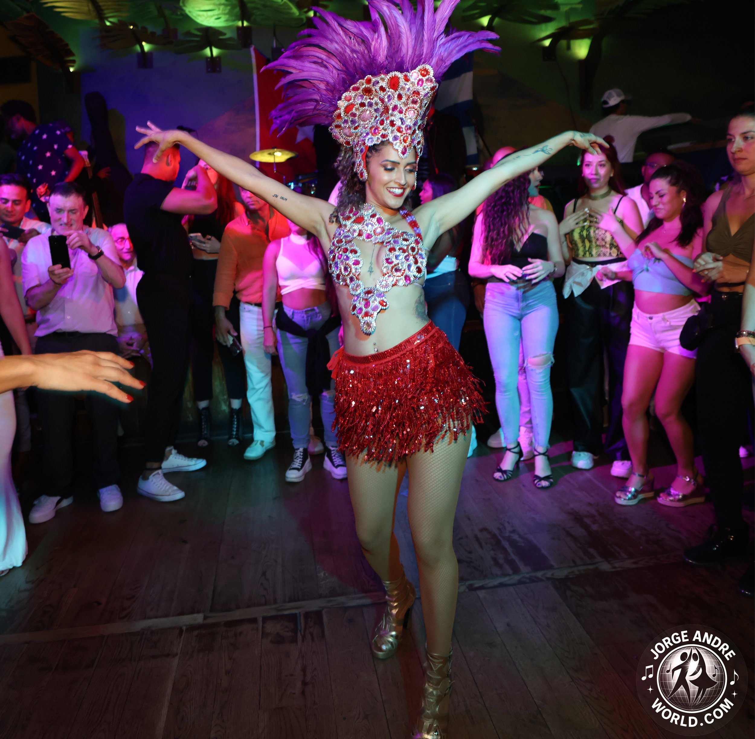 A woman in a colorful, elaborate costume with feathers, sequins, and gemstones is dancing joyfully in the middle of a crowd at a lively party or celebration.