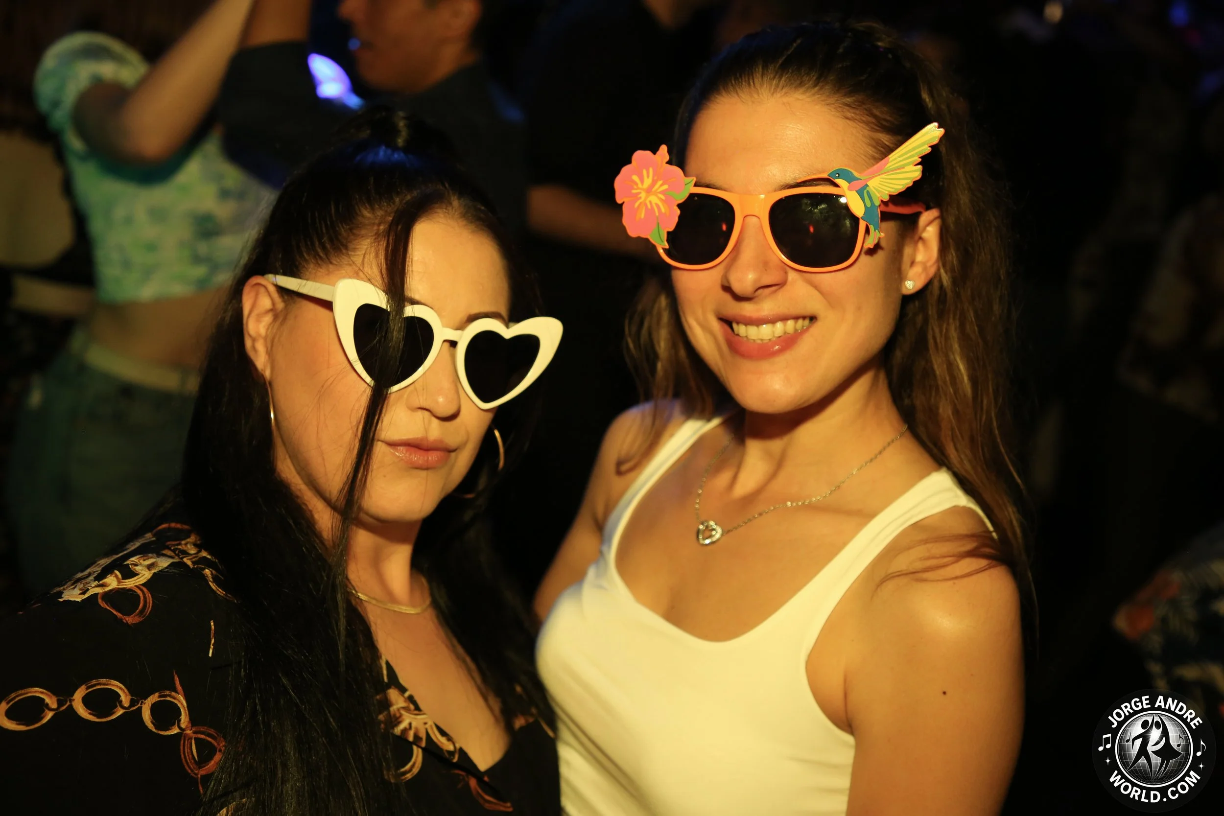 Two women at a party wearing colorful sunglasses with decorations; one has heart-shaped sunglasses and the other has glasses with a flower and hummingbird design, both smiling.