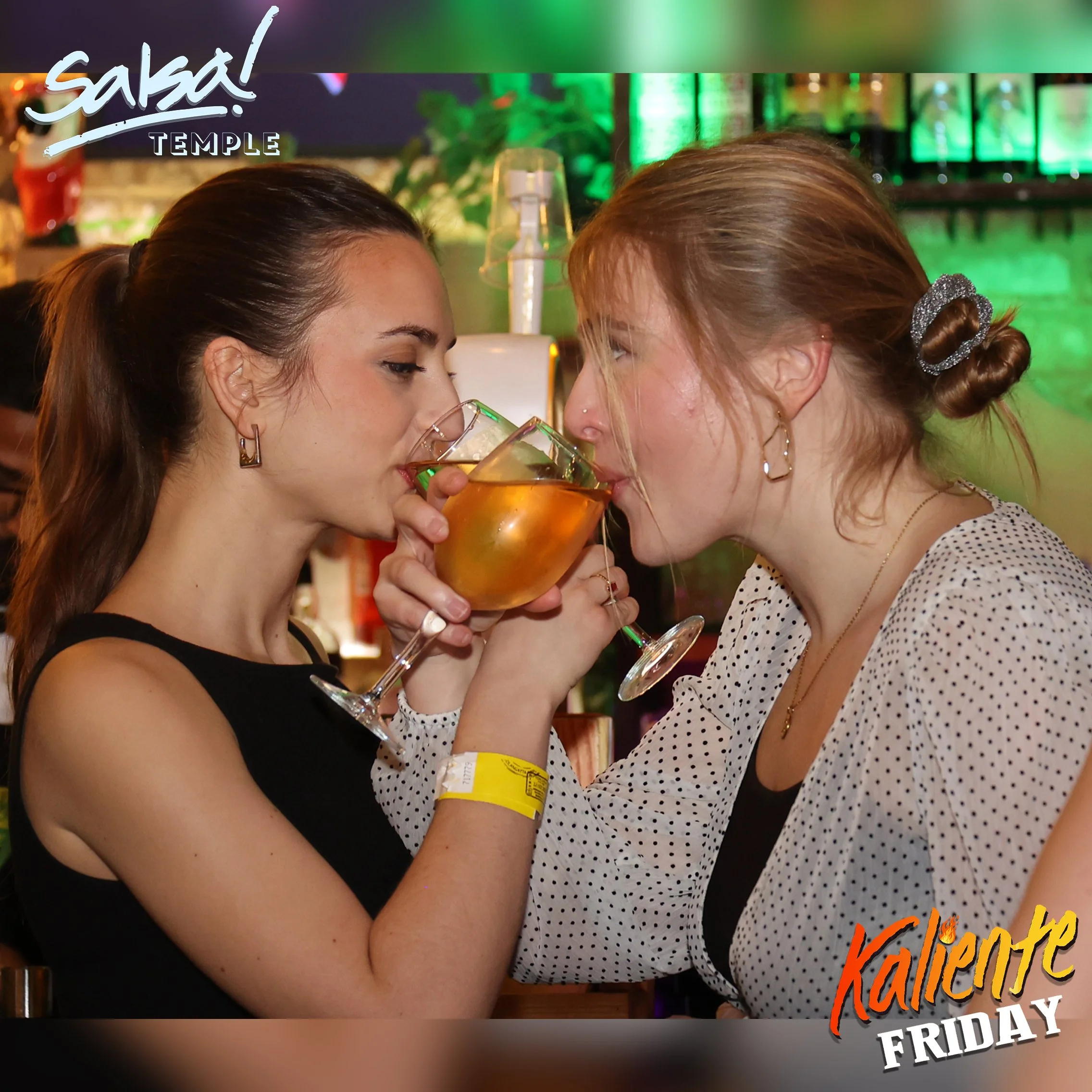 Two women clinking glasses with drinks at a lively bar or restaurant, celebrating Friday night.