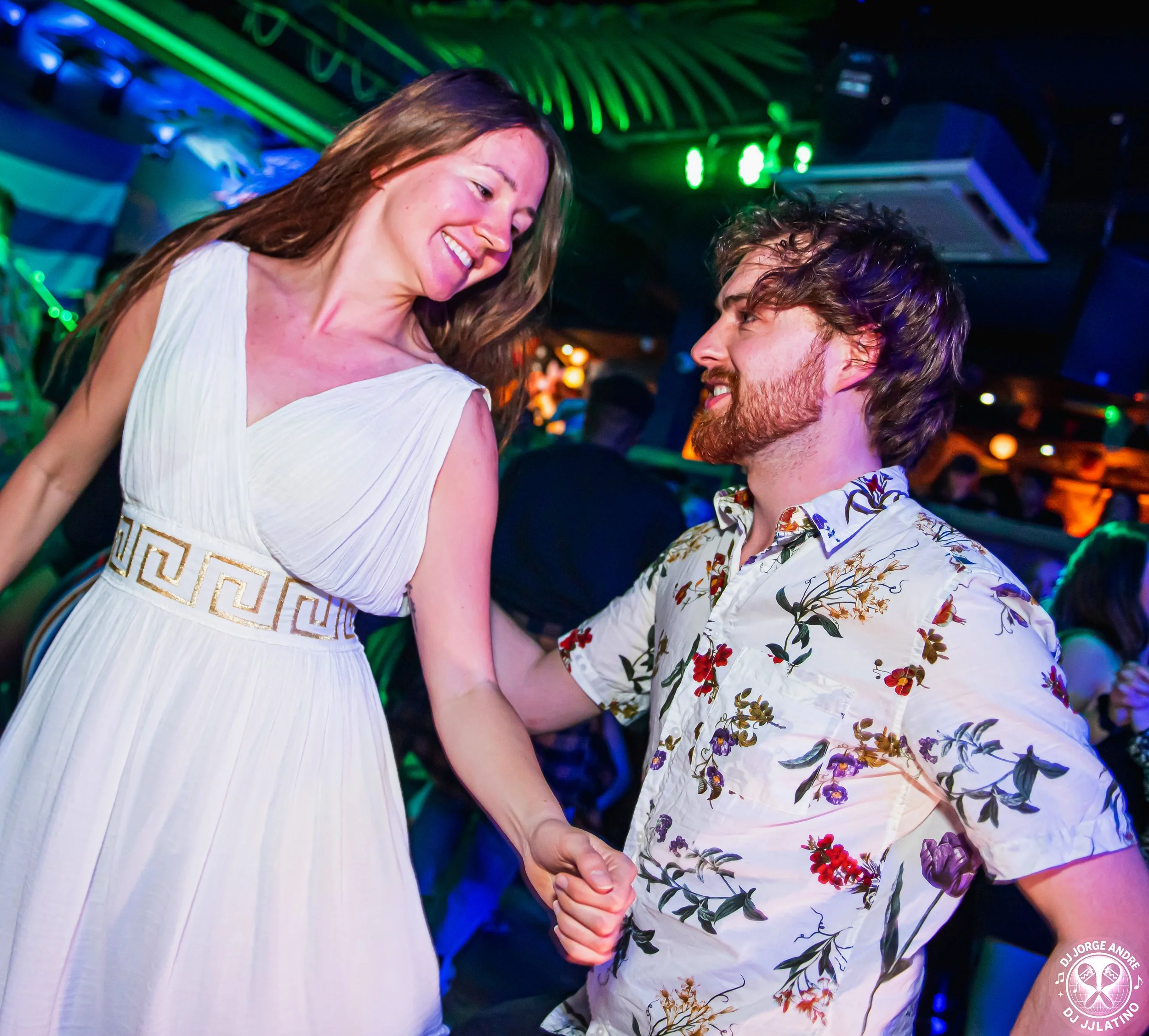 A woman and a man dancing together at a lively, colorful party or nightclub, smiling and holding hands.
