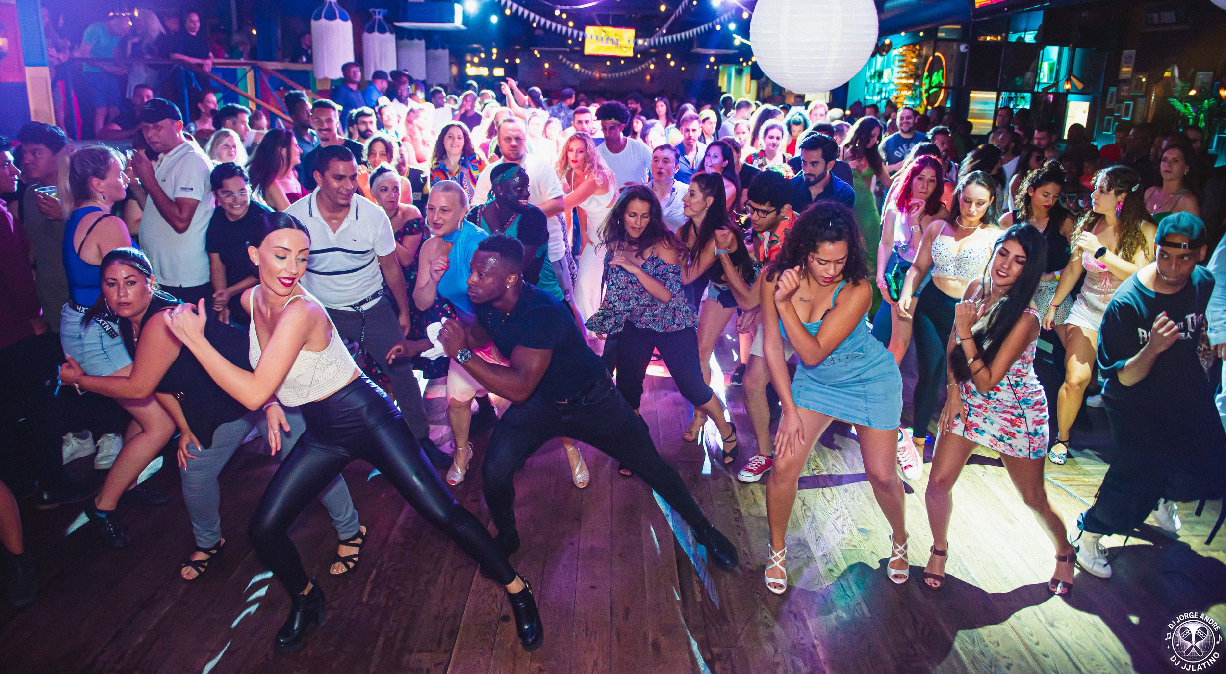 Crowd of people dancing at a lively party or nightclub with colorful lighting and decorations.