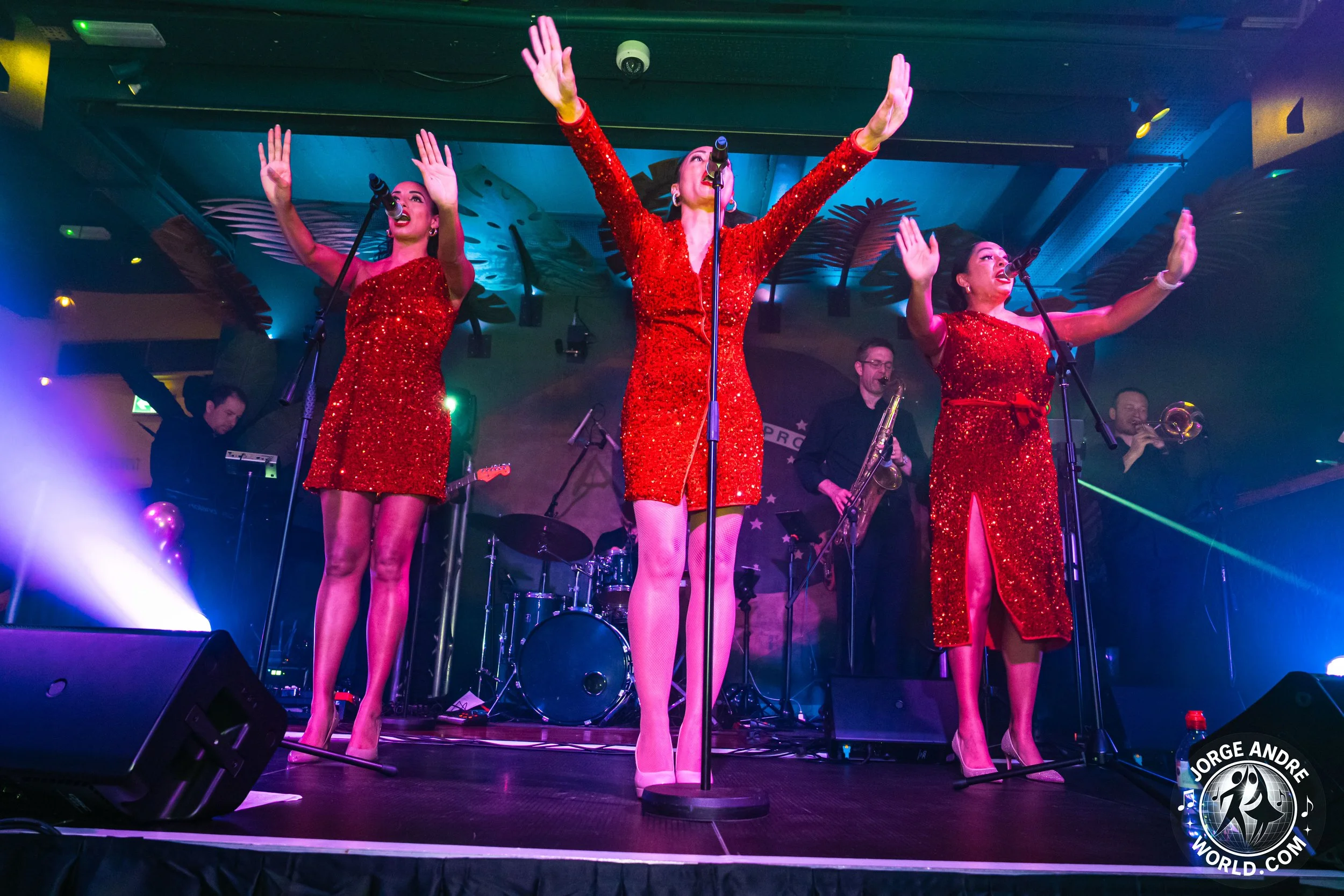 Three female singers in red sequined dresses with high slits on stage, singing with their arms raised, accompanied by a band with musicians playing saxophone, trumpet, keyboard, and drums, under colorful stage lighting.