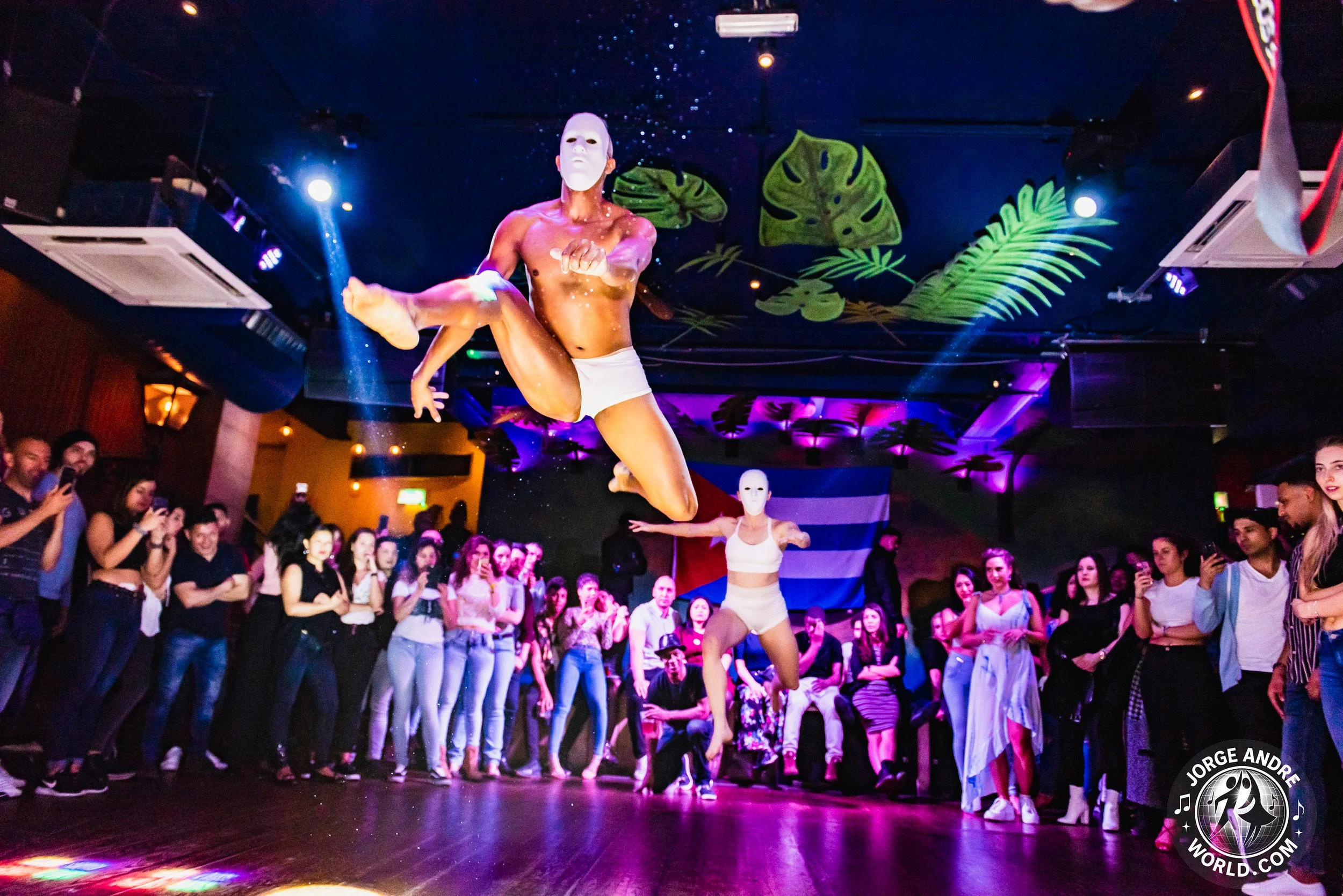 People watching a dance performance on a dance floor, with two performers wearing white masks and minimal clothing, one mid-air leap, colorful lighting, tropical leaf decorations on the ceiling, and a Greek flag in the background.