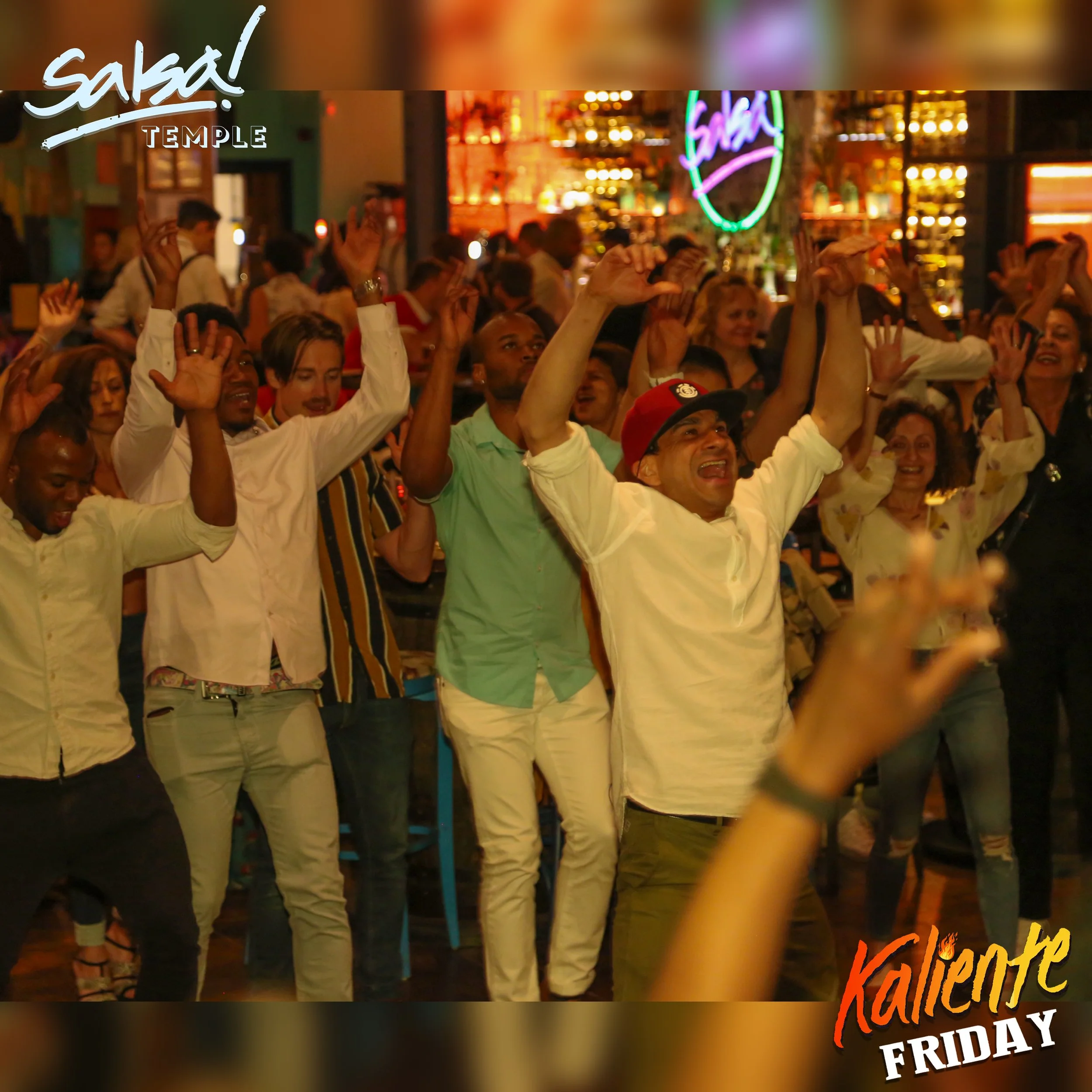 A group of people dancing and celebrating in a lively indoor setting with colorful lights and a neon sign that says "Salsa." The atmosphere suggests a salsa dance event or party, with diverse attendees enjoying the music and dance.