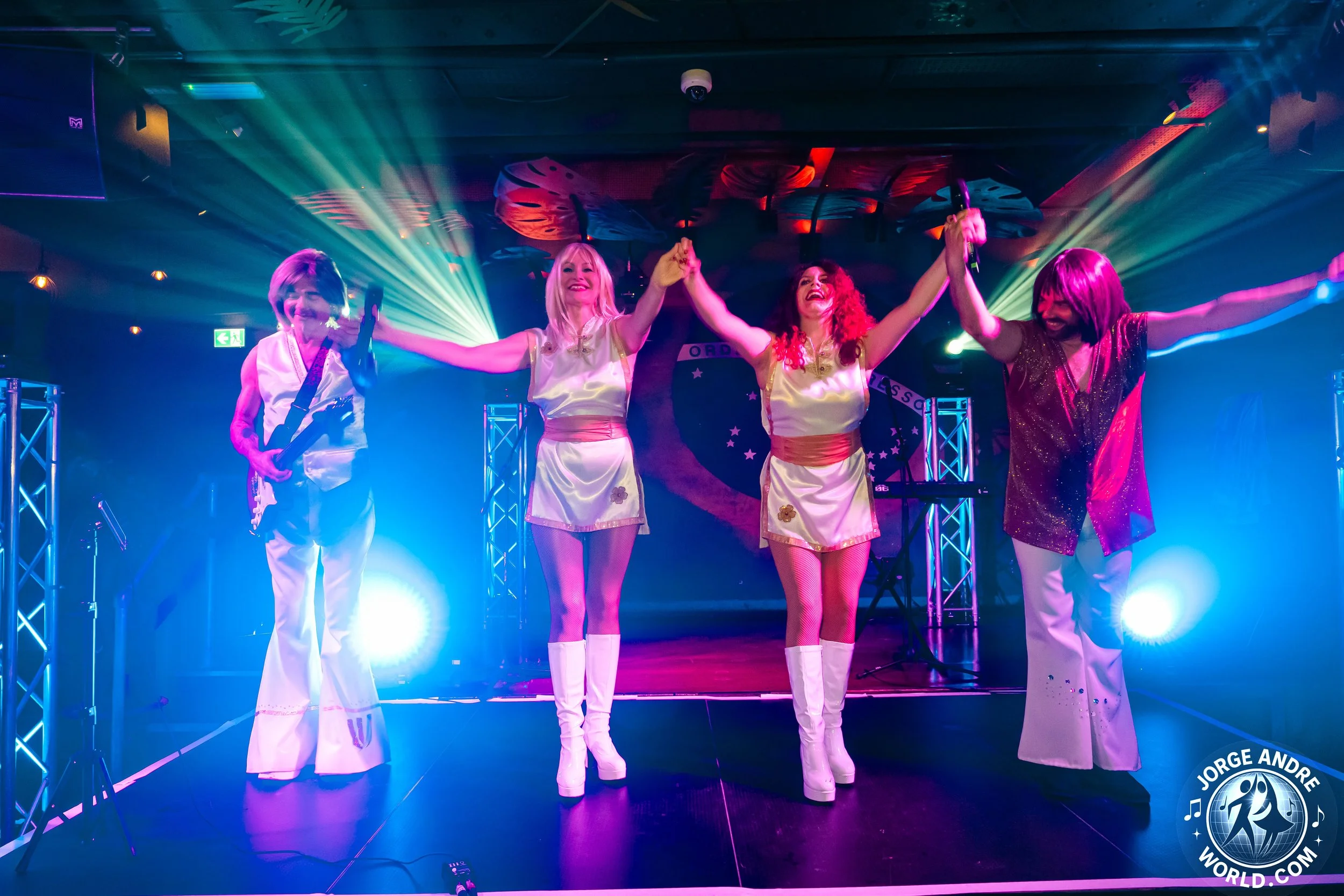 Four women on stage at a concert, holding hands with arms raised, smiling, with colorful stage lights shining behind them, and stage equipment visible.