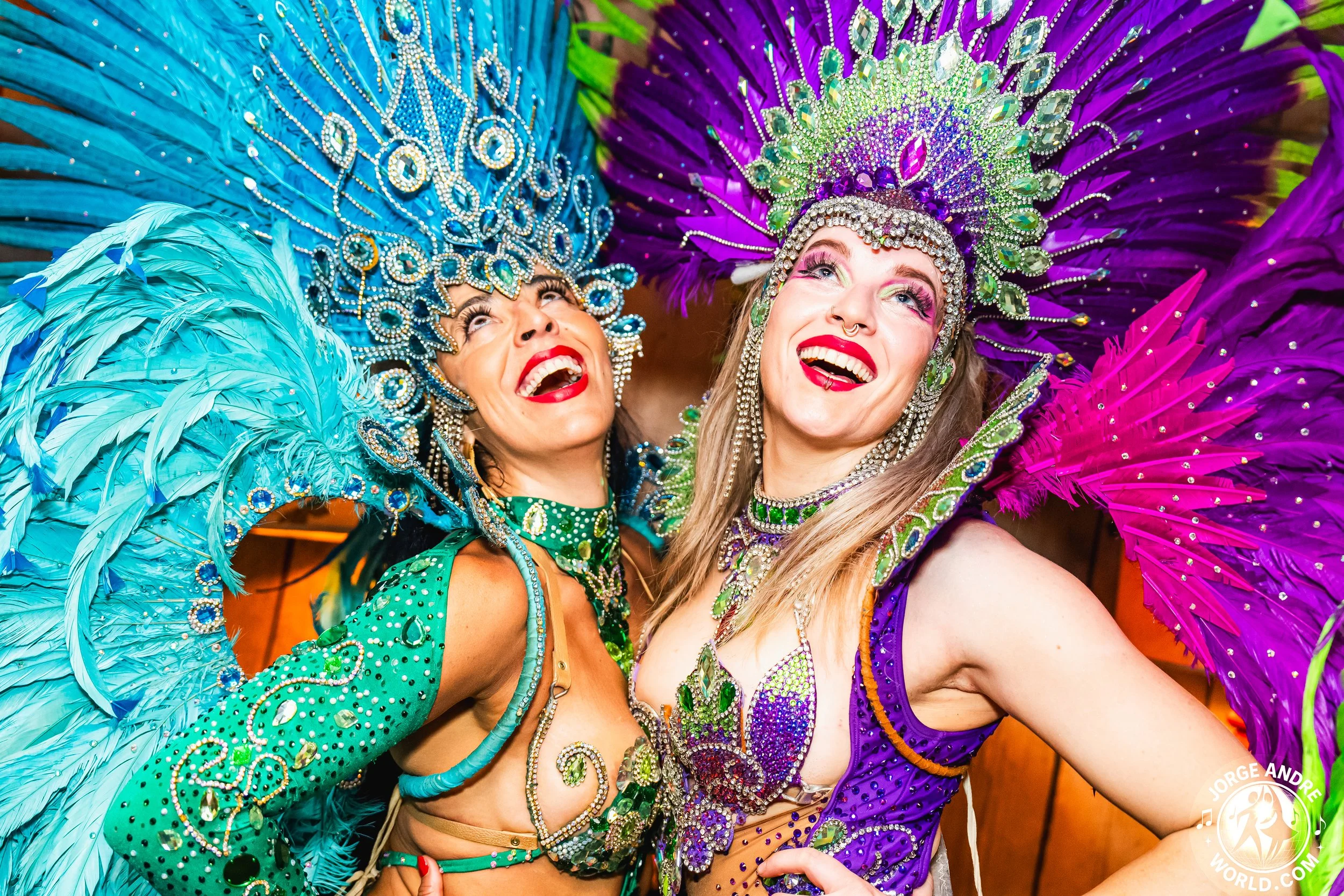 SAMBA DANCERS, Two women dressed in colorful, elaborate carnival costumes with feathered headdresses and rhinestone embellishments, smiling and looking joyful.