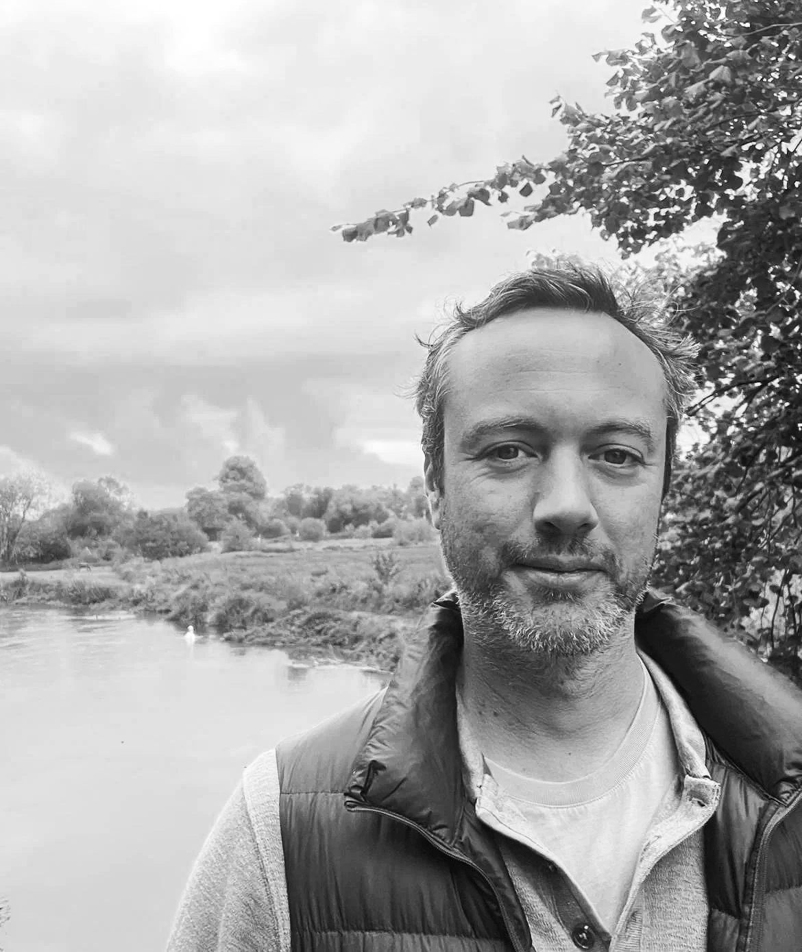 A black-and-white photo of a Alexander Gray, standing outdoors near a river, with trees and shrubs in the background and cloudy sky overhead.