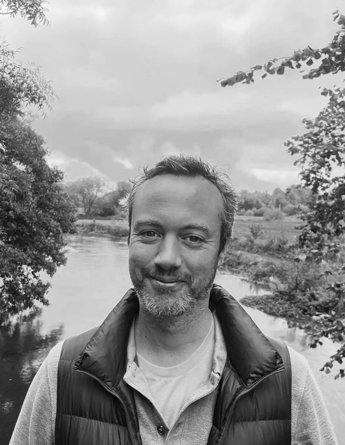 Black and white photo of Alexander Gray, principal therapist at Garden of Eadon, standing outdoors near a river with trees and a cloudy sky in the background.