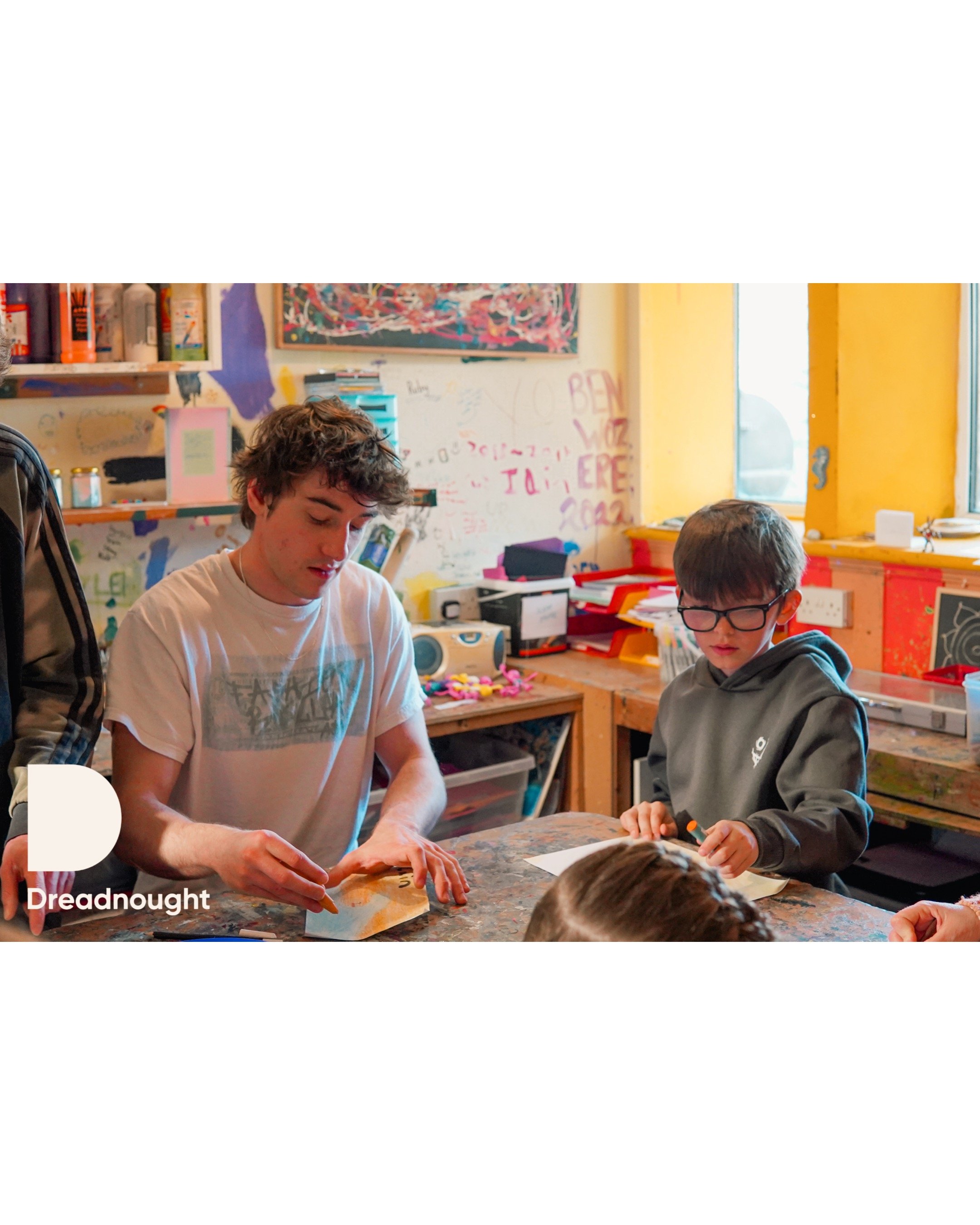 Two young boys sitting at a table in a colorful, graffiti-decorated room with shelves of art supplies and a window, engaged in an art project.