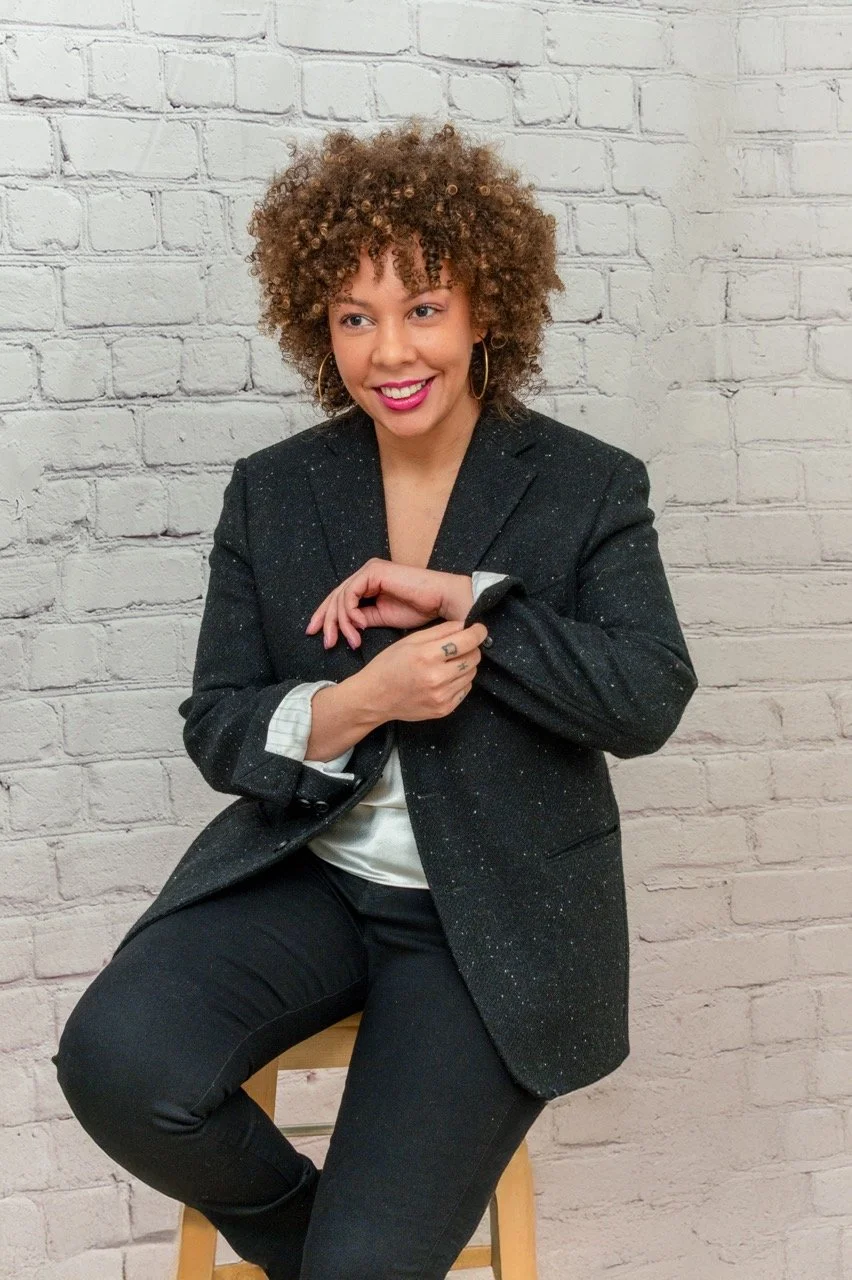 The Owner Raia with curly hair and hoop earrings sitting on a wooden stool, wearing a black blazer and white shirt, smiling against a white brick wall.