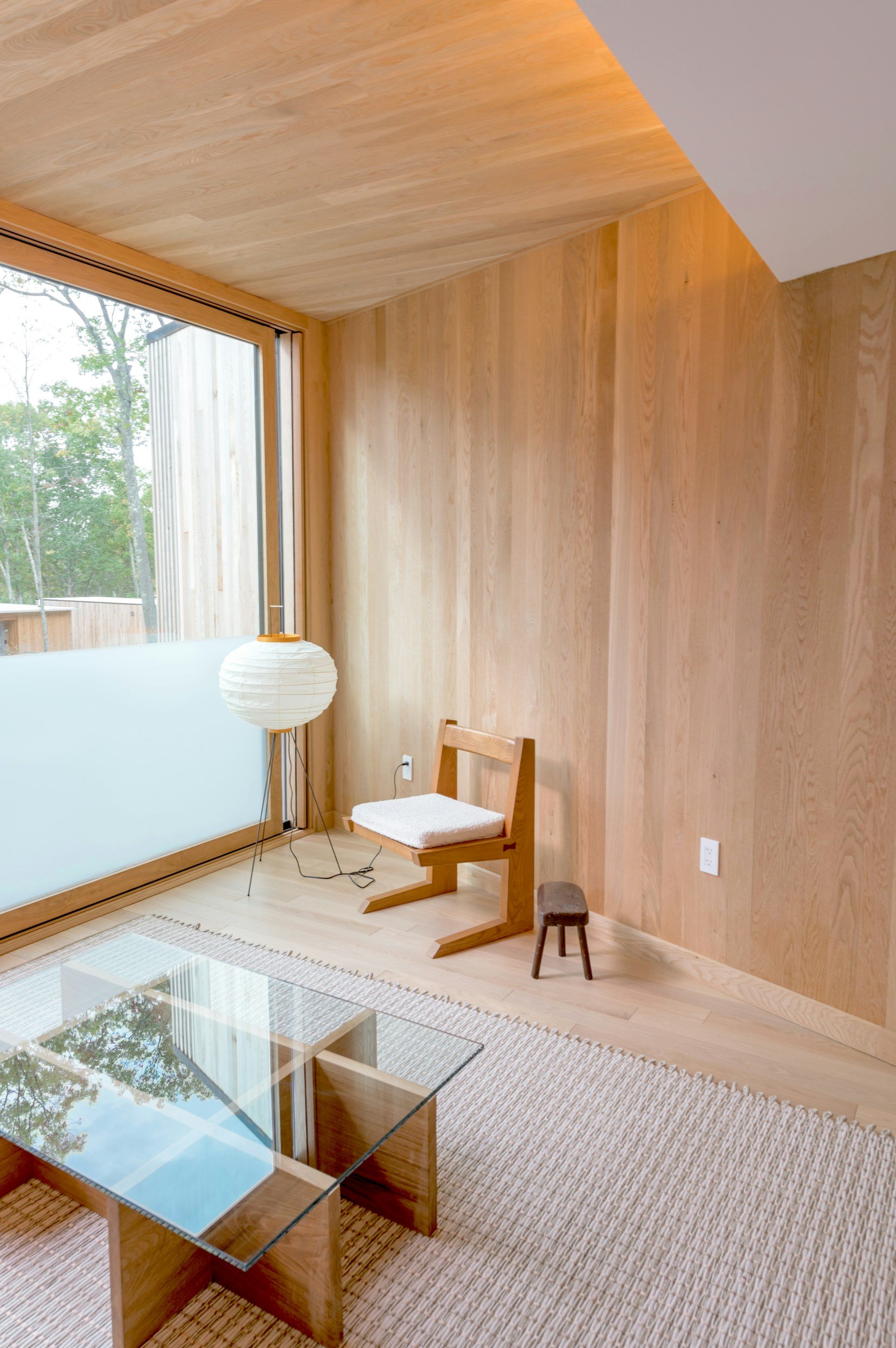 A wooden interior room with large window, wooden walls and ceiling, a white paper lantern lamp, a wooden chair with white cushion, a small wooden stool, a striped rug, and glass coffee table visible in the foreground.