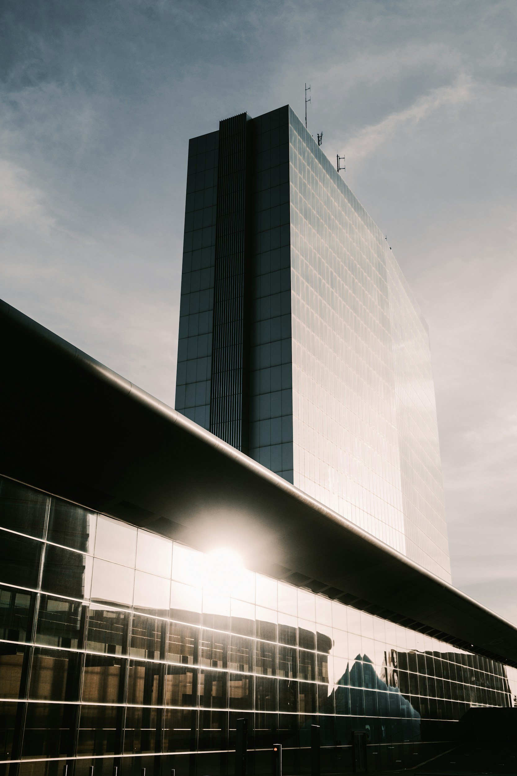 Picture of a tall modern office building in Kirchberg, Luxembourg, with a glass facade reflecting the sky, seen from an angle with part of the lower structure also visible.