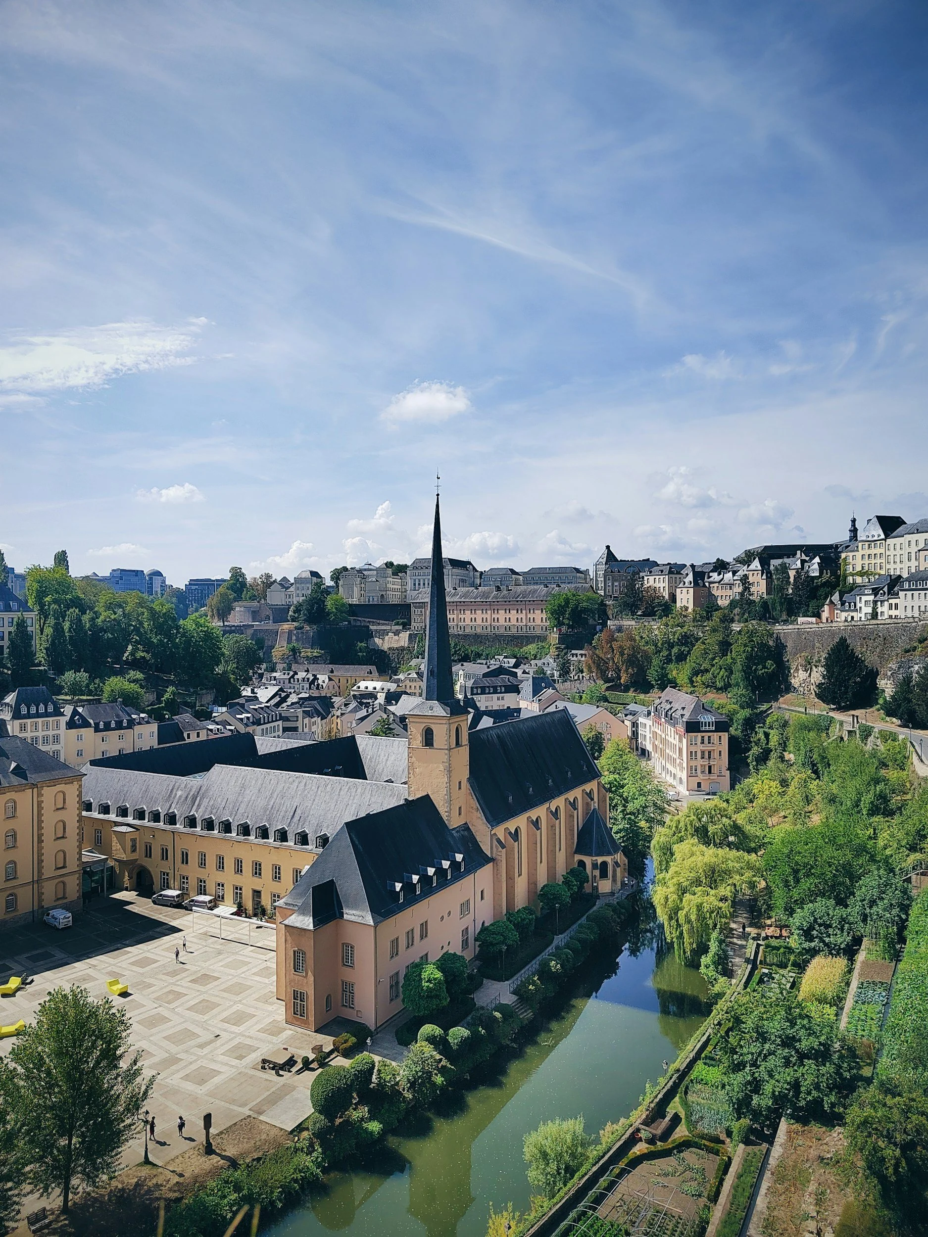 Picture of Luxembourg city, featuring a church with a tall steeple, surrounded by historic buildings, a river with greenish water, and lush green trees under a partly cloudy blue sky.