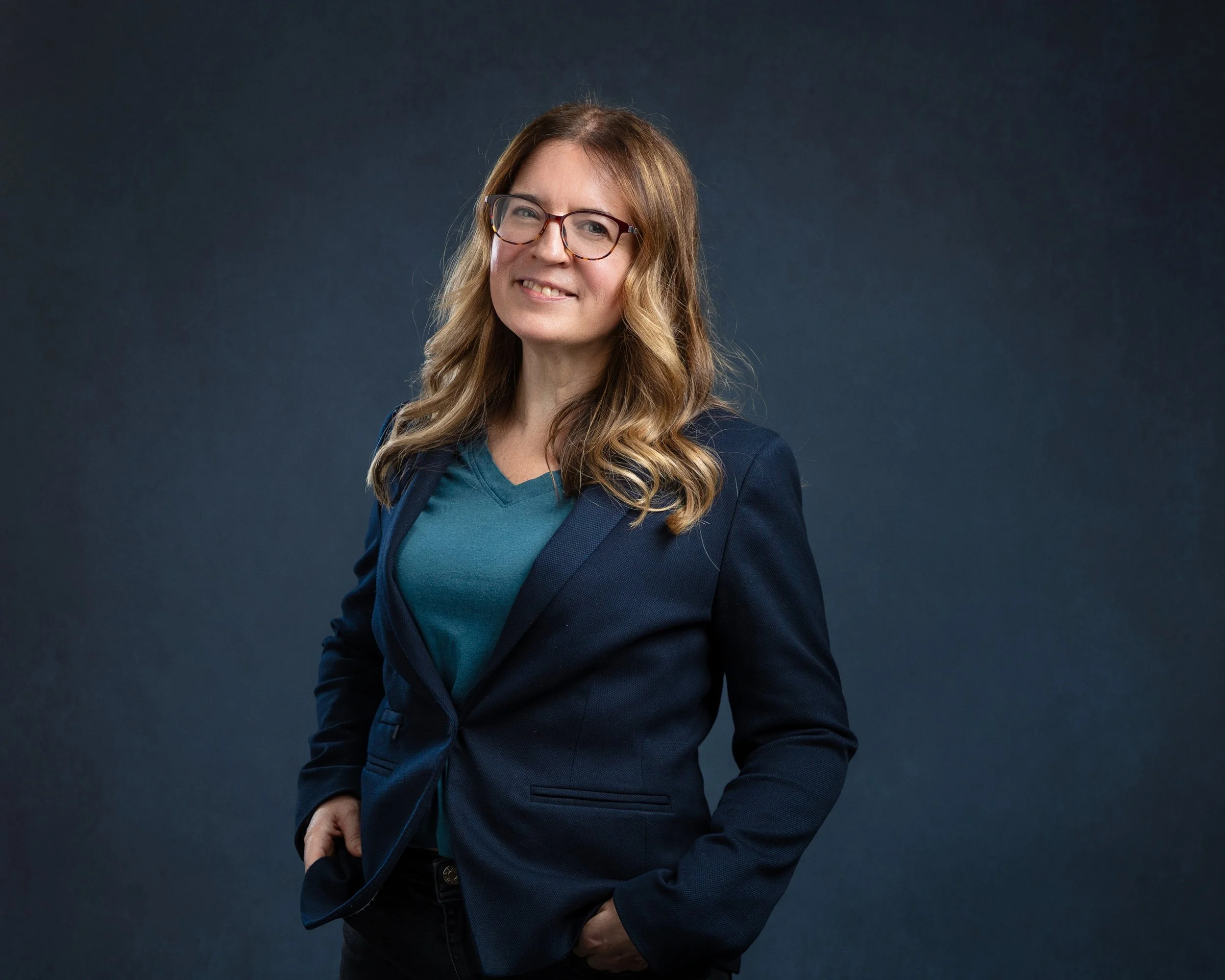 Picture of Yvonne Reilly, with long wavy blonde hair, glasses, wearing a dark blazer over a teal shirt, standing against a dark gradient background.