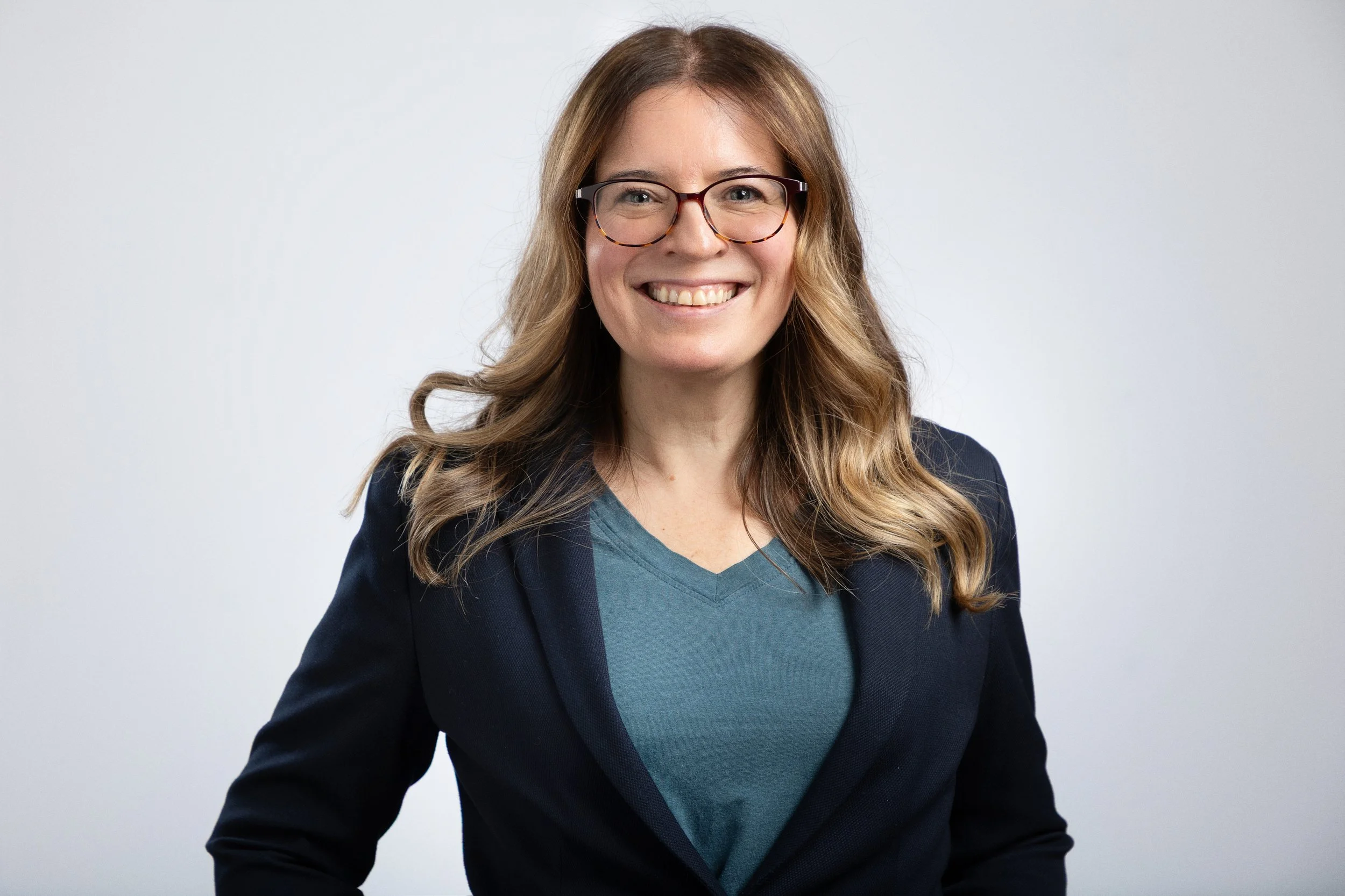 Picture of Yvonne Reilly, a smiling woman with light brown wavy hair, wearing glasses, a blue V-neck shirt, and a black blazer, standing against a plain light gray background.