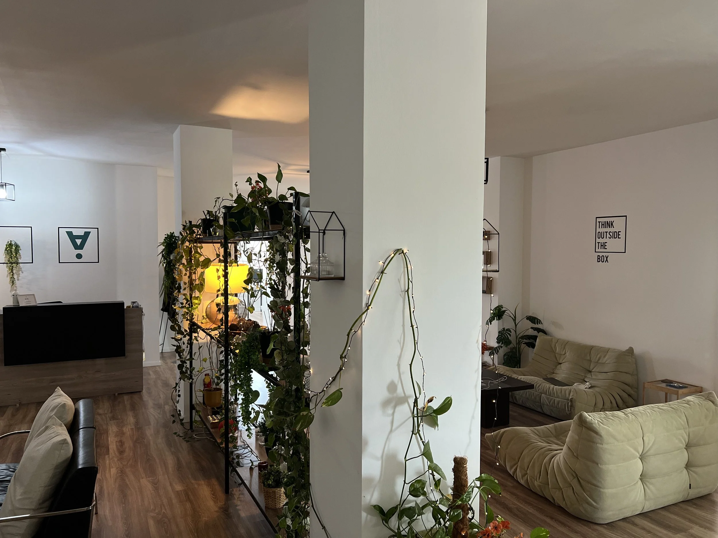 A cozy living space with two beige sofas, a black coffee table, and a wooden side table. Decorative lights and potted plants are present, with wall art including a framed quote, 'Think outside the box,' and framed symbols.