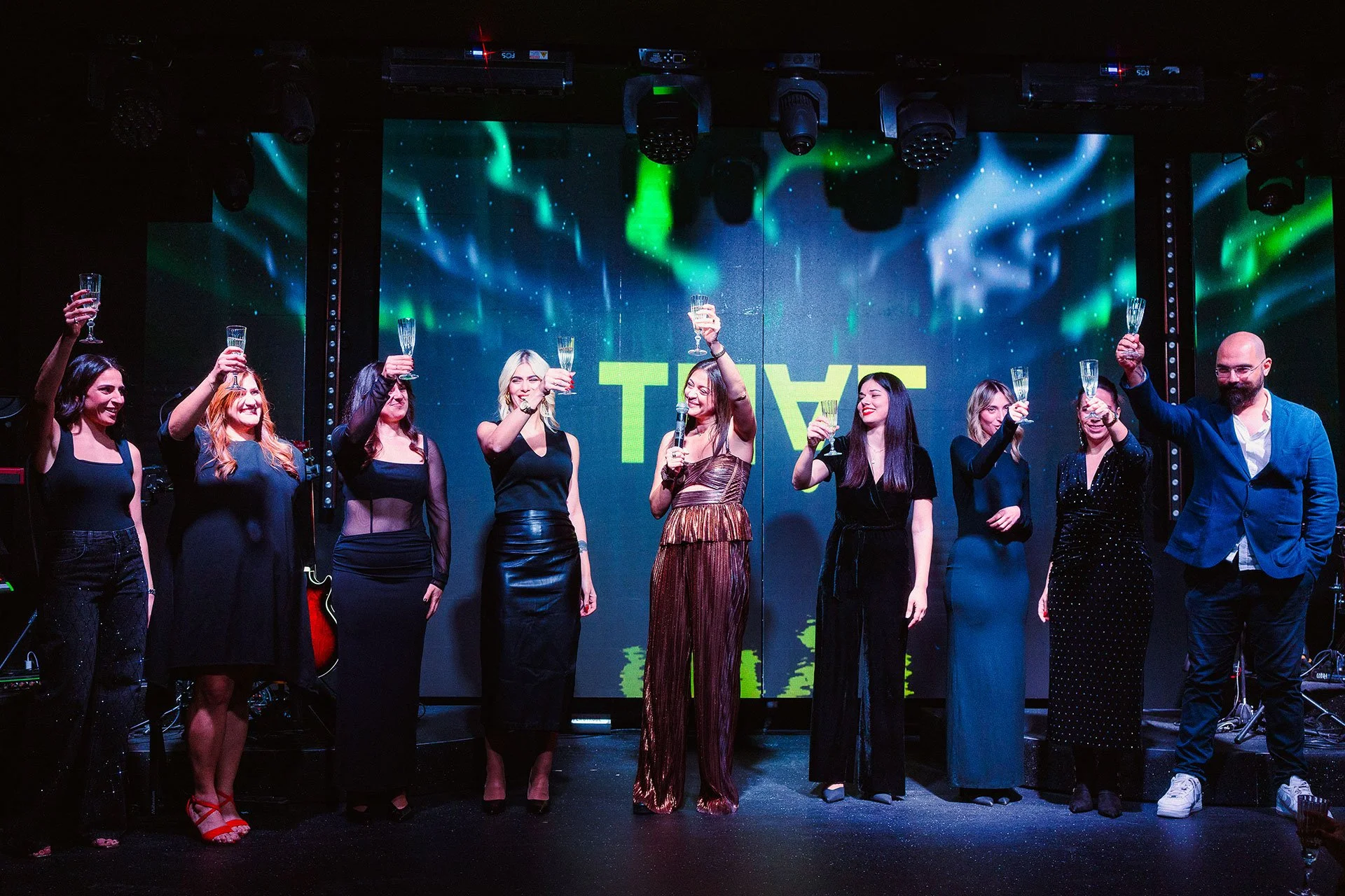 Group of nine people on stage celebrating with toasts at a formal event, background with colorful lights and large screen displaying text.