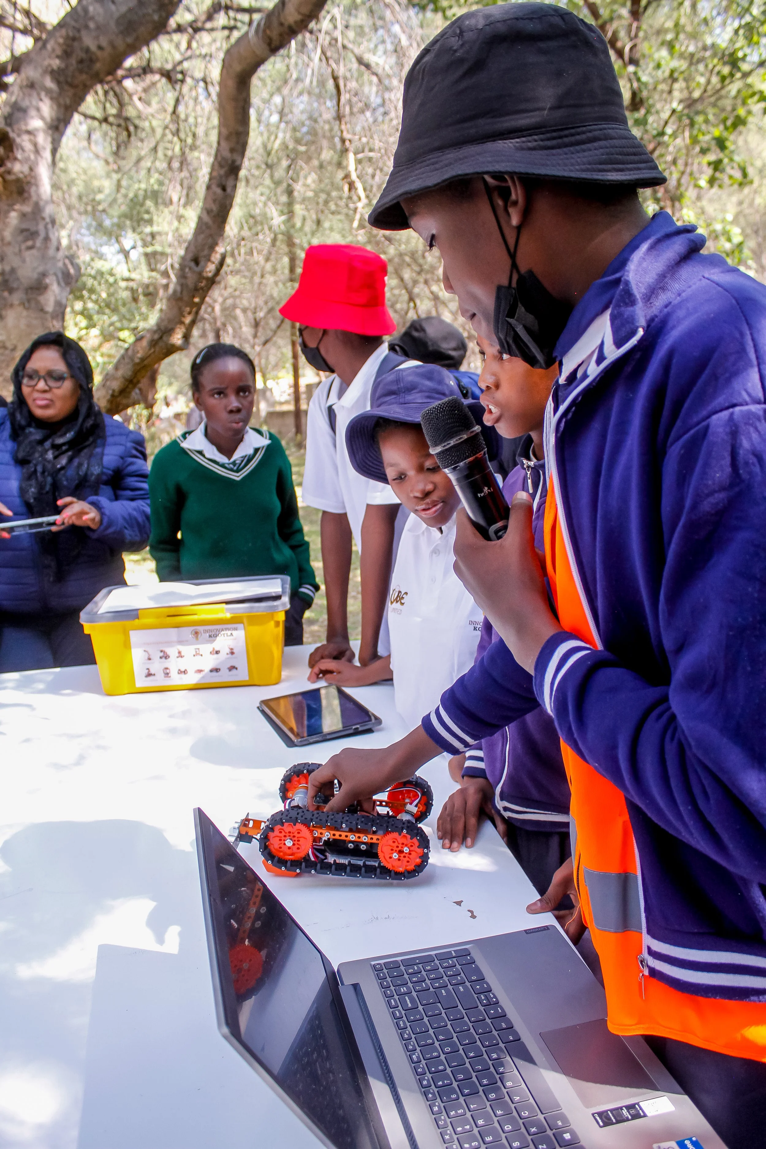 A group of children and adults outdoors, gathered around a table, observing a small robotic vehicle. One child is holding a microphone, explaining the robot to the group. On the table, there's a laptop, a tablet, and a yellow box with labels.