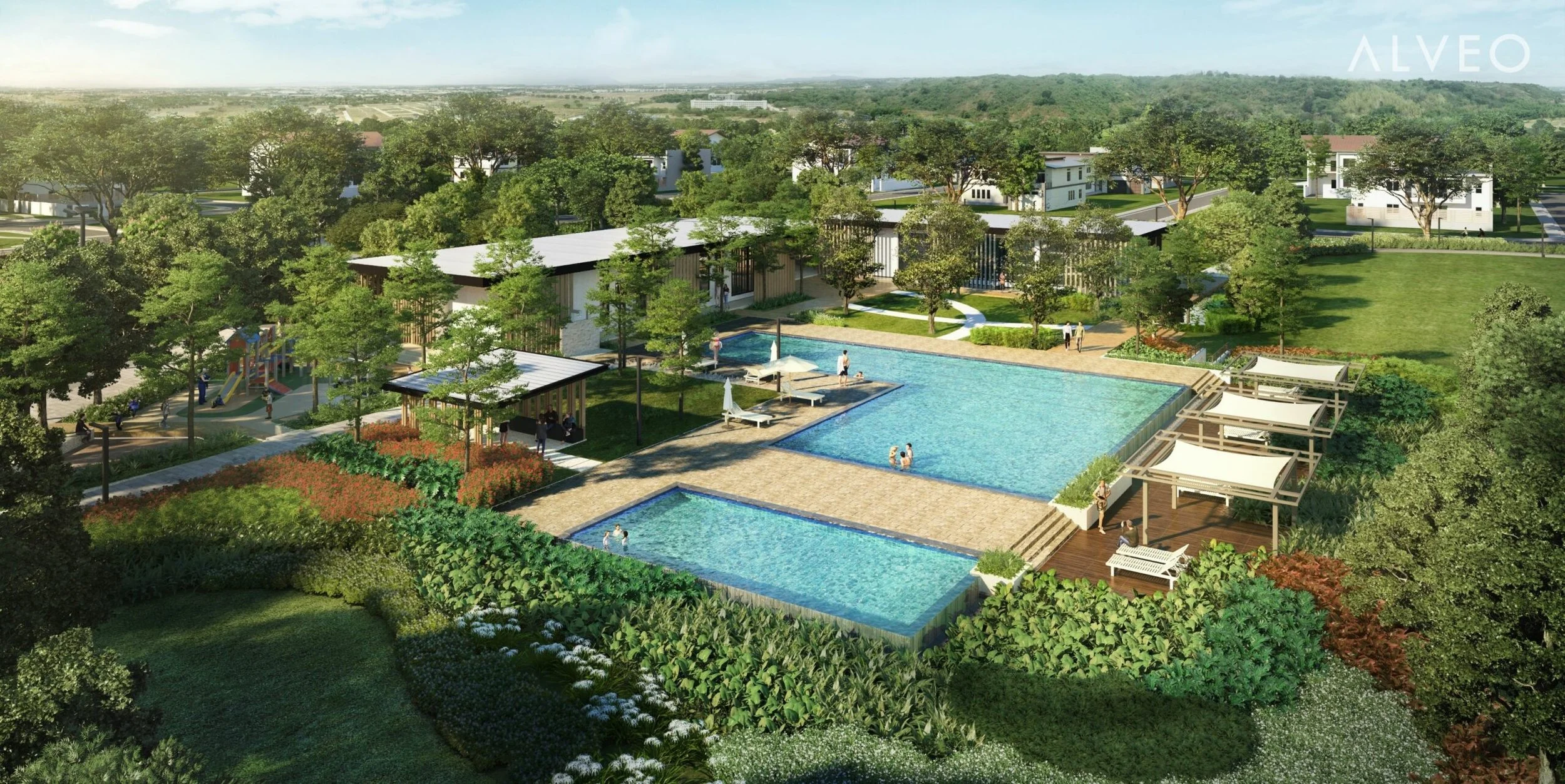 Aerial view of a residential complex with multiple swimming pools, surrounded by green lawns, trees, and walking paths, with houses and hills in the background.