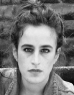 Portrait en noir et blanc d'une femme avec des cheveux bouclés et un regard intense, posant devant un mur en pierre.