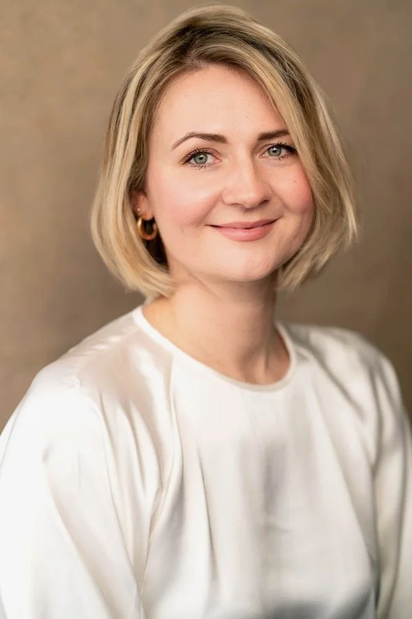 A woman with blonde, shoulder-length hair, blue eyes, and light makeup, smiling slightly while wearing gold hoop earrings and a white satin blouse, standing against a plain beige background.