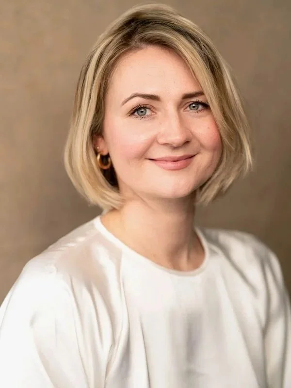 A woman with blonde hair wearing a white top, smiling, with a neutral background.