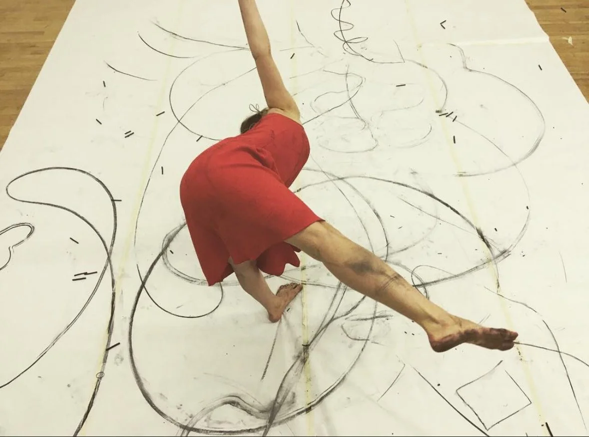 Person in red shorts performing a handstand on an abstract, large-scale drawing on the floor with black lines and shapes.