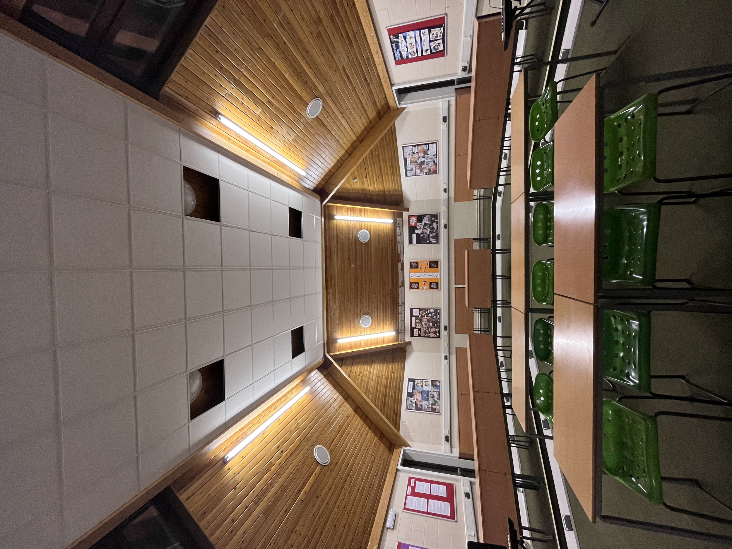Indoor classroom or meeting room with wooden ceiling, fluorescent lighting, tables, green chairs, bulletin boards, and wall posters.