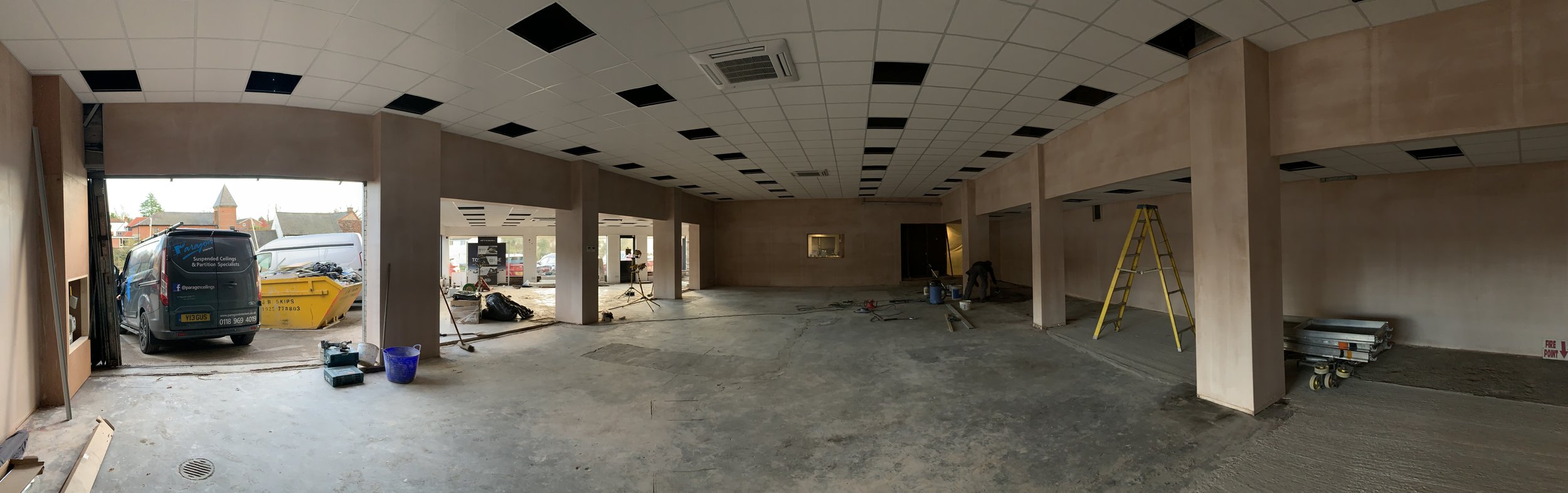 Interior of a building under construction or renovation with open garage-style door, construction tools, ladder, and construction waste visible.