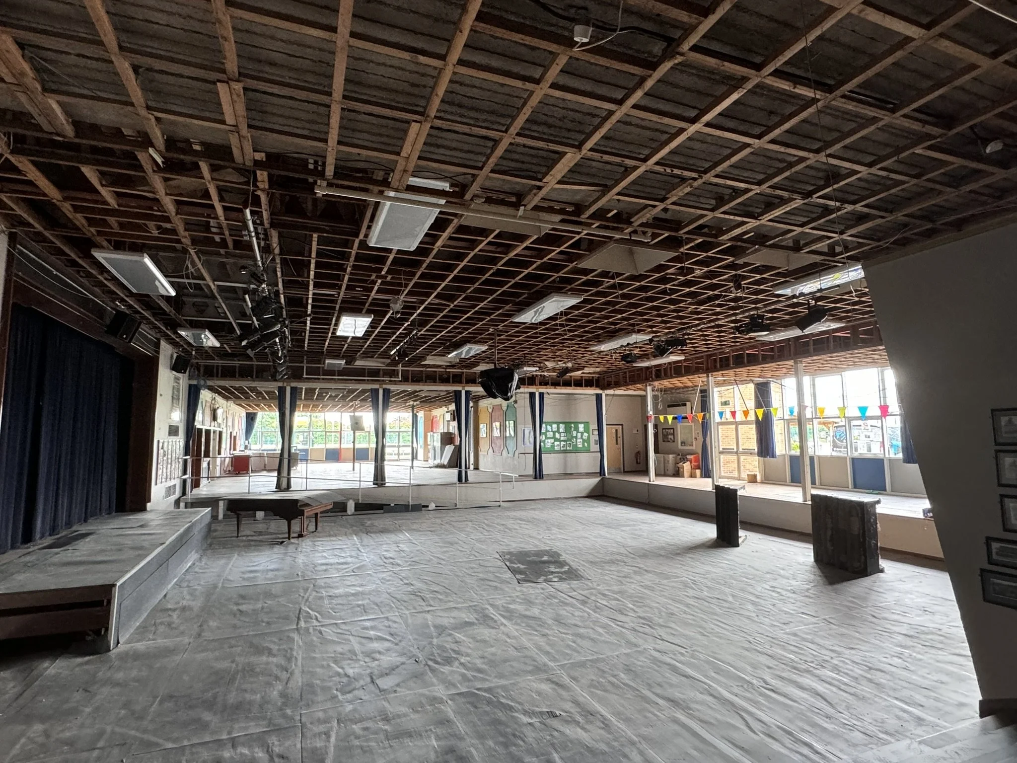Interior of a large, empty room with a high, exposed wooden grid ceiling and several suspended fluorescent light fixtures. The floor appears to be covered in a tarp or protective material, and there are stage steps on the left side. The room has larg