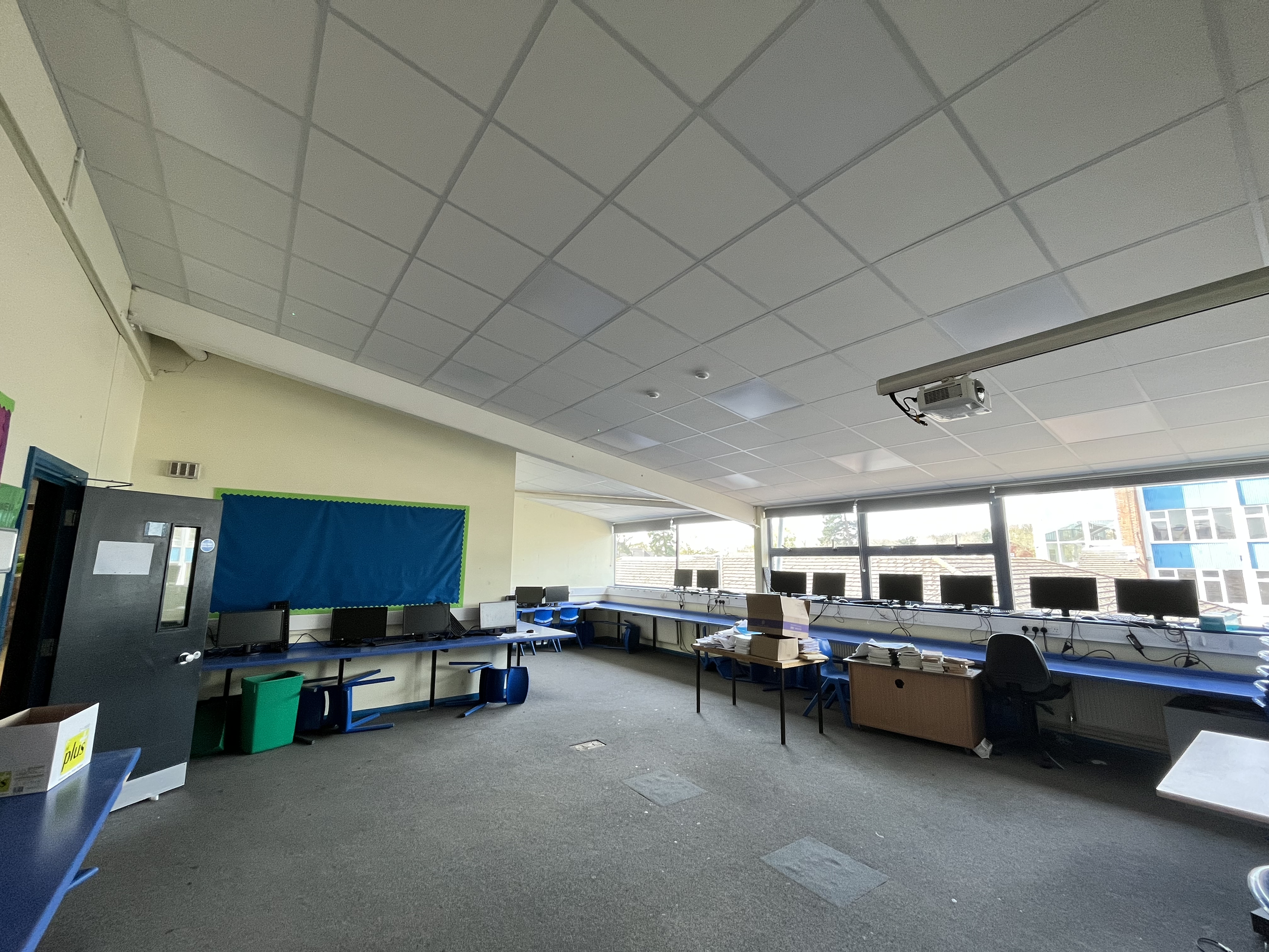 An empty classroom with desks and computers along the windows, ceiling lights, and a projector mounted on the ceiling.
