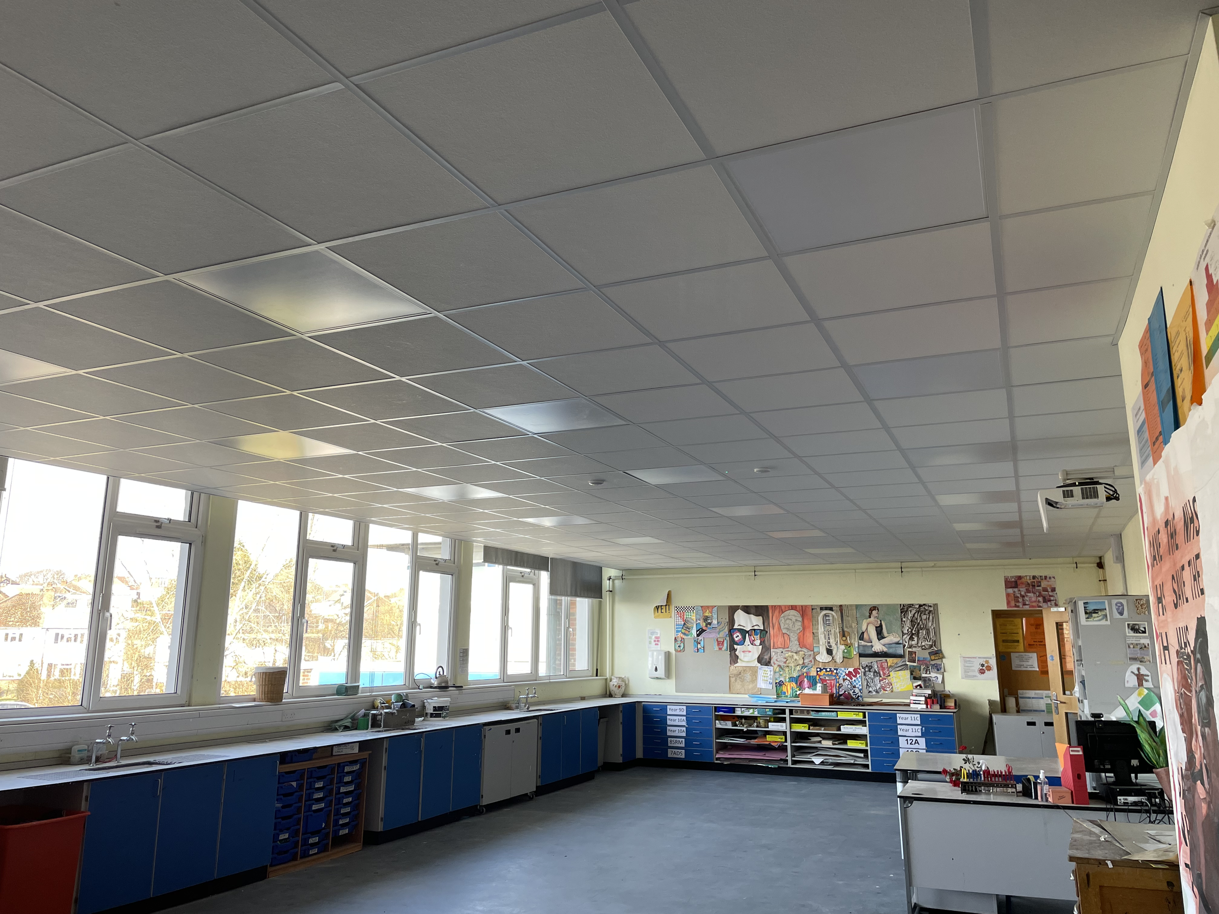 Classroom with large windows, blue storage cabinets, colorful artwork on the wall, and a ceiling with grid-patterned tiles.