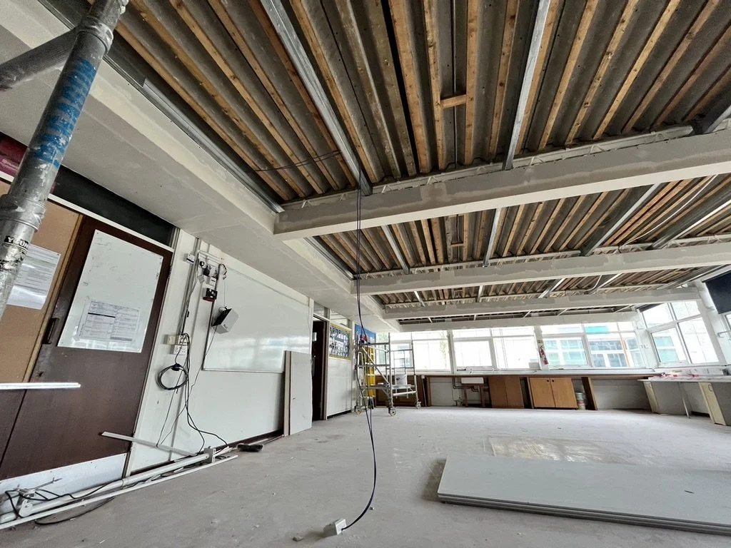 Under construction or renovation in a room with exposed ceiling beams, construction tools, and materials like drywall on the floor, large windows, and some cabinetry.