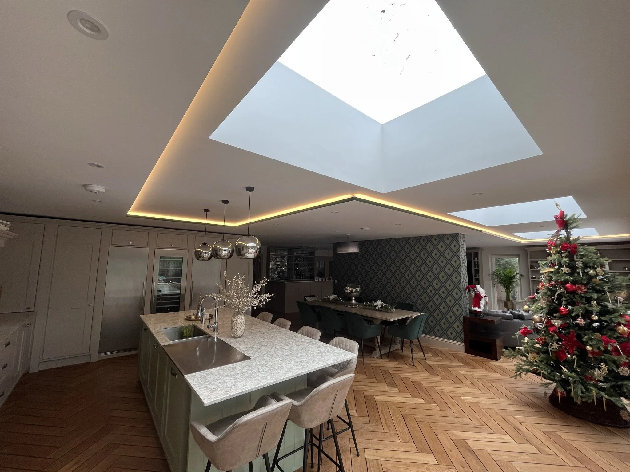 View of a modern kitchen and dining area with a large skylight in the ceiling, decorated for Christmas with a decorated Christmas tree on the right side, and a table with chairs in the foreground.