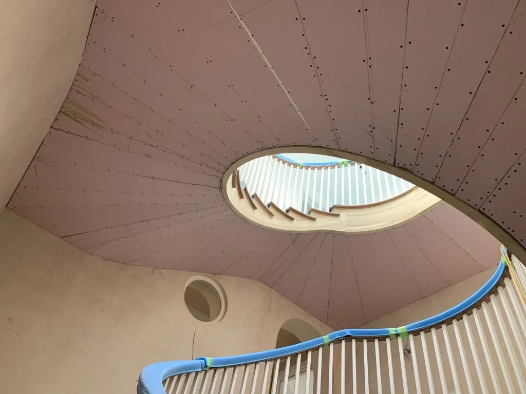 Interior view of a spiral staircase looking up through an ovoid opening in a pink ceiling with a curved blue handrail.