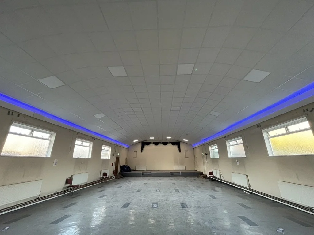 Empty large hall with a stage, multiple windows, and blue accent lighting along the ceiling.