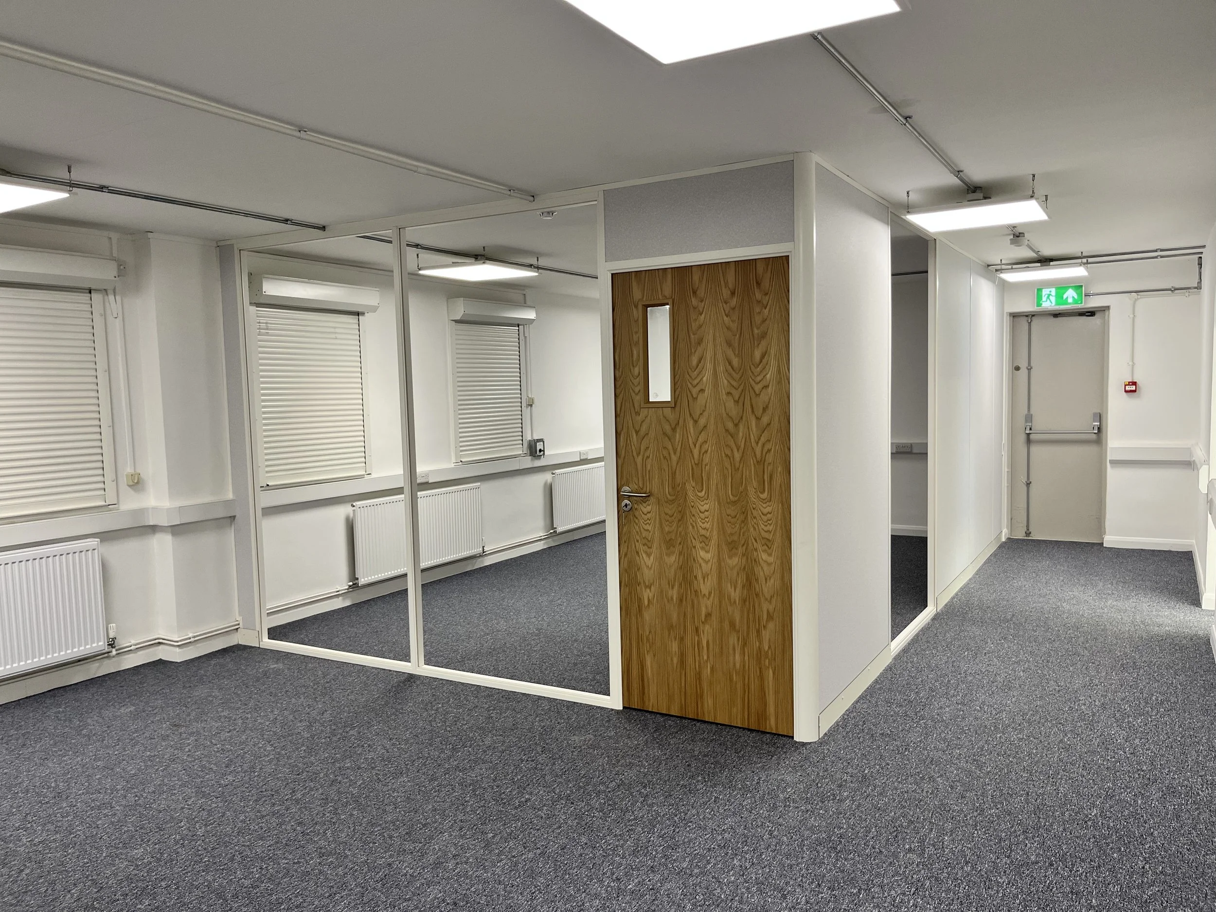 Empty office space with white walls, gray carpet flooring, and a small enclosed booth with wood paneling.