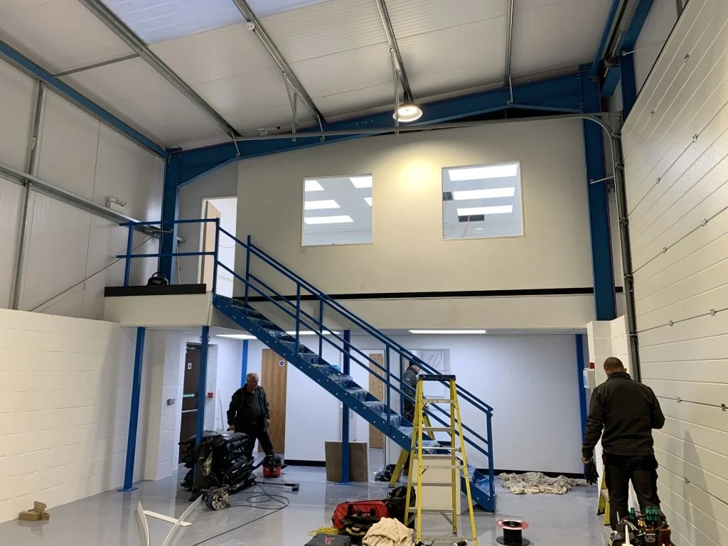 Interior of a warehouse or industrial building under construction, with a blue staircase leading to a second-floor balcony, two workers, construction tools, and materials.