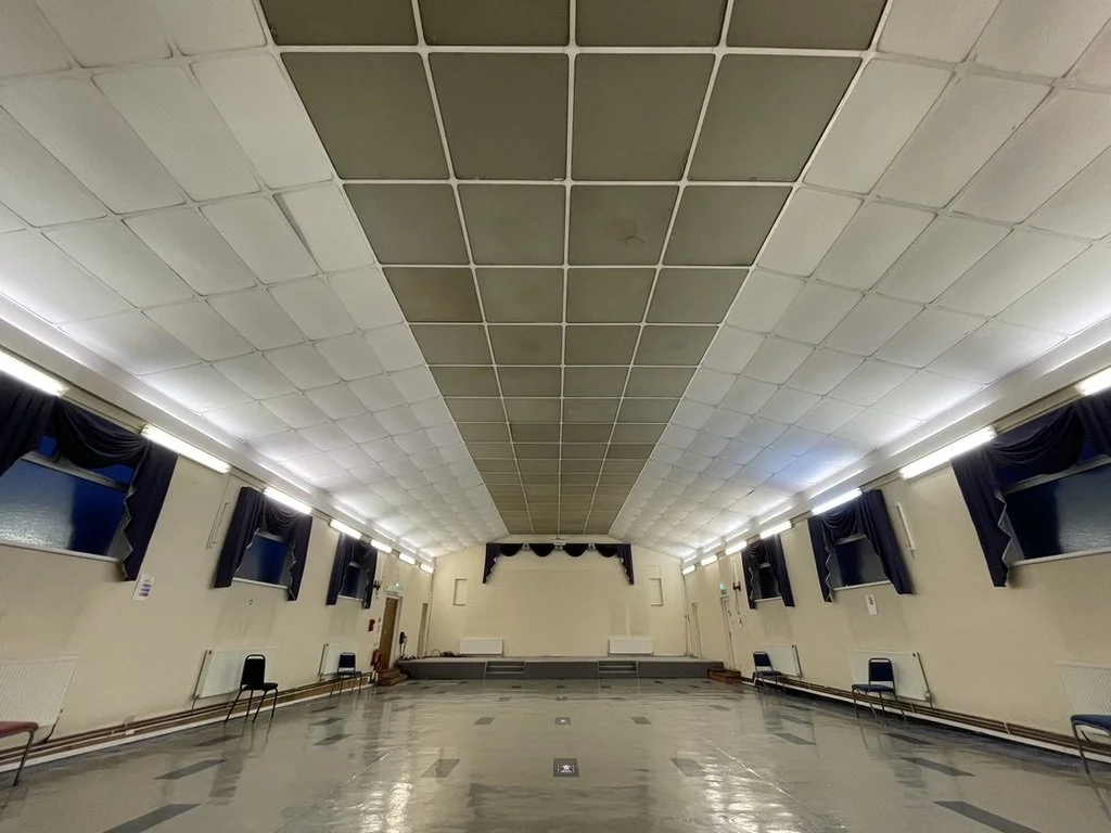 Empty community hall or event room with chairs along the walls, windows with dark curtains, and a stage at the front. The ceiling has a large section of patterned tiles.