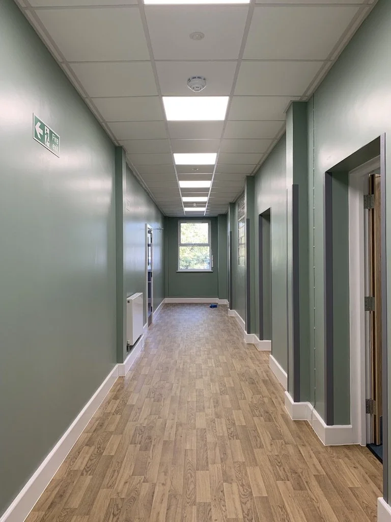 Empty corridor in a building with green walls, wooden floor, ceiling lights, and multiple doors on the right side. Window at the end of the hall showing trees outside.