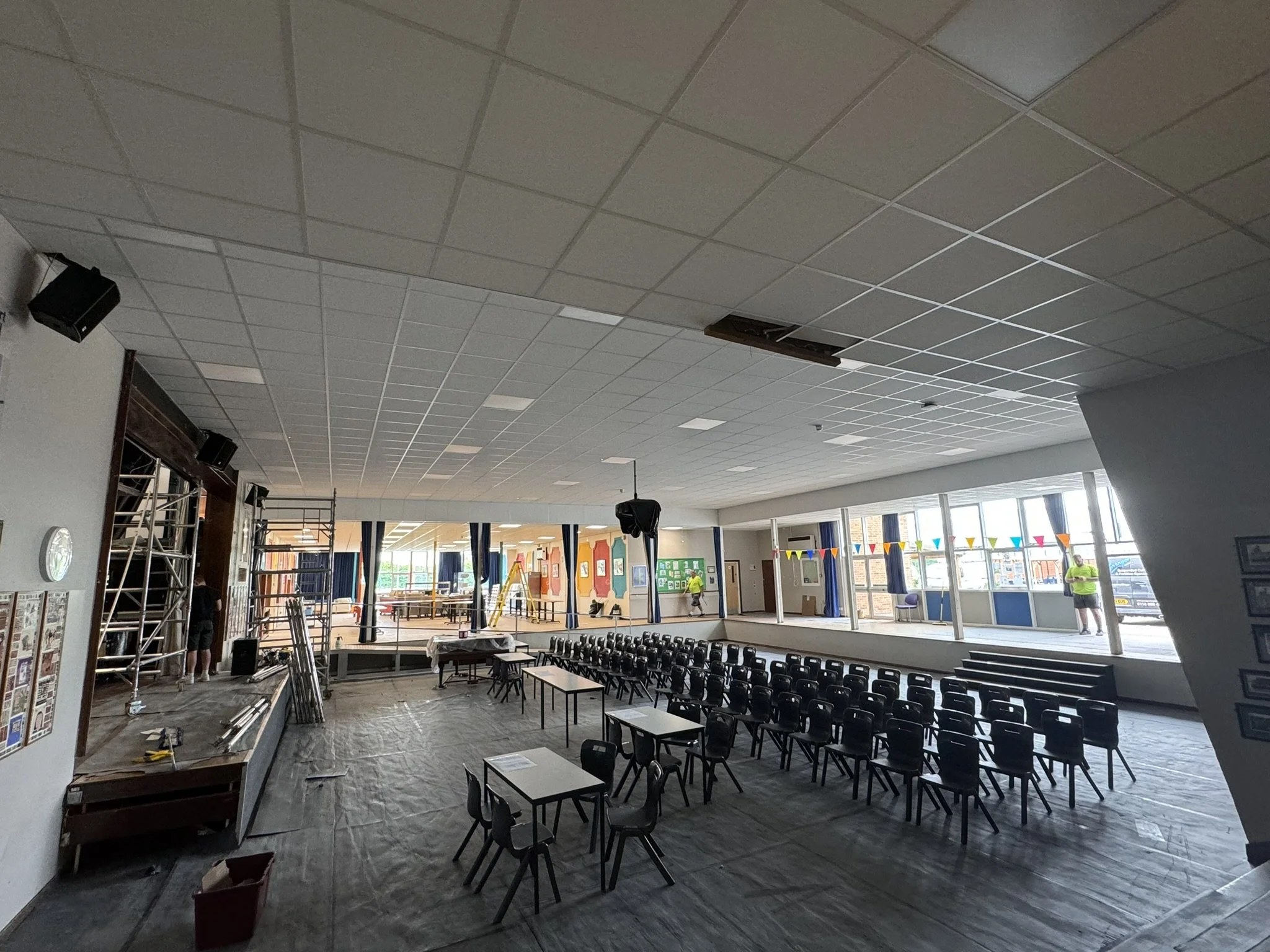 Interior of a large room, possibly a gymnasium or auditorium, with a high ceiling covered in square tiles. The room is in the process of being set up or renovated, with scaffolding, ladders, and construction materials visible. Rows of black chairs ar